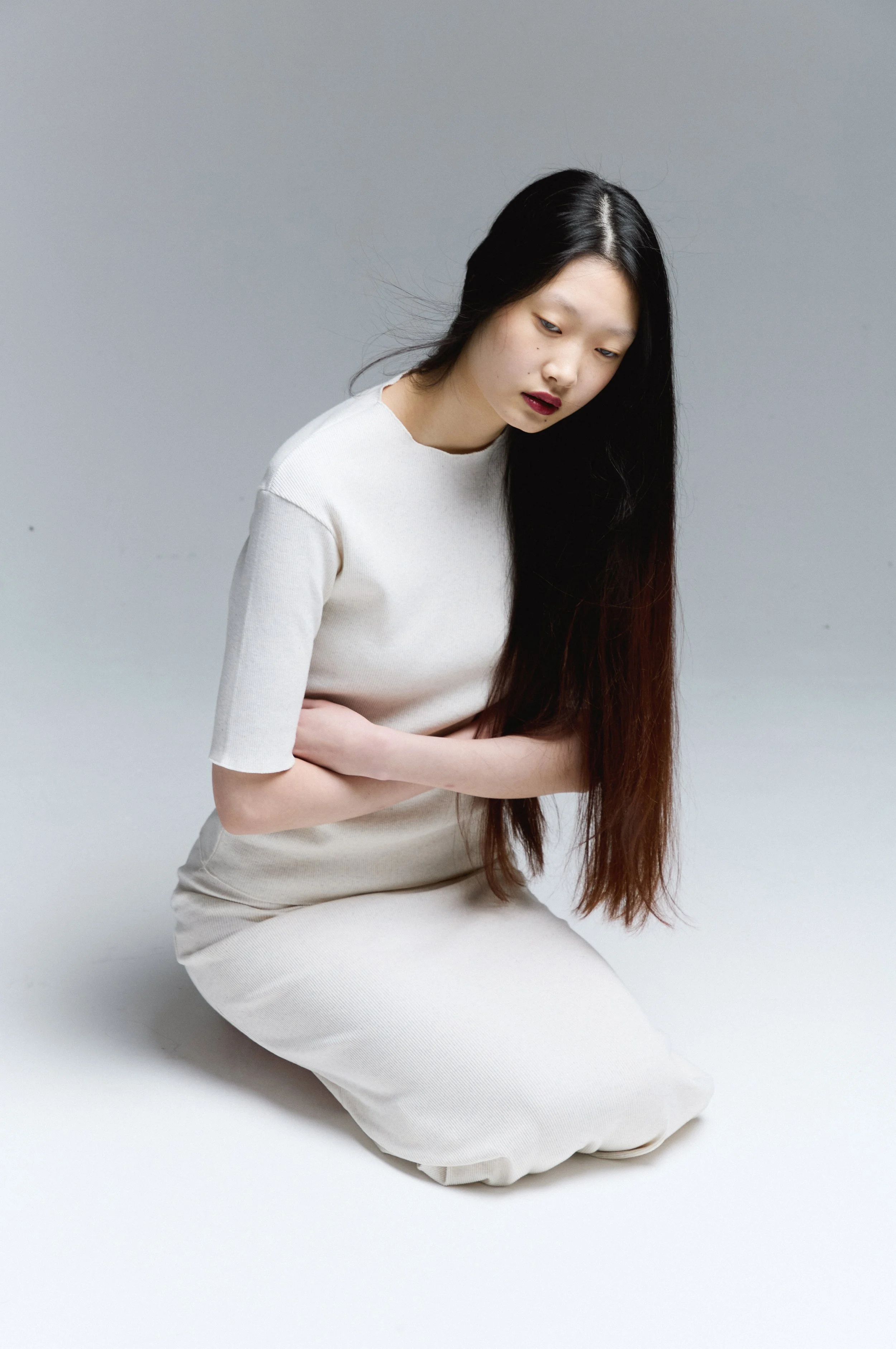 Young woman with long black hair, wearing a cream-colored dress, sitting in a kneeling pose against a plain gray background, with her head tilted downward.