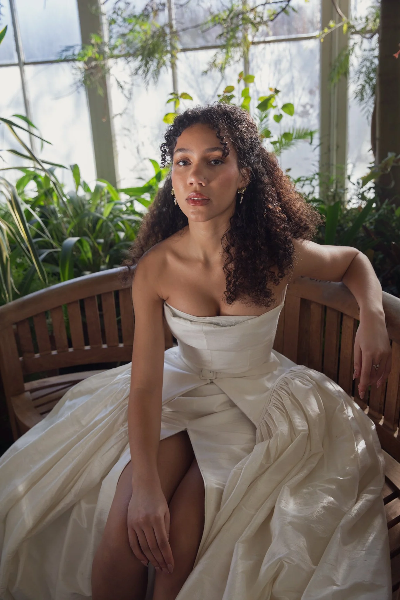 A woman with curly hair and earrings sitting on a wooden bench surrounded by plants, wearing a strapless dress, inside a greenhouse or conservatory.