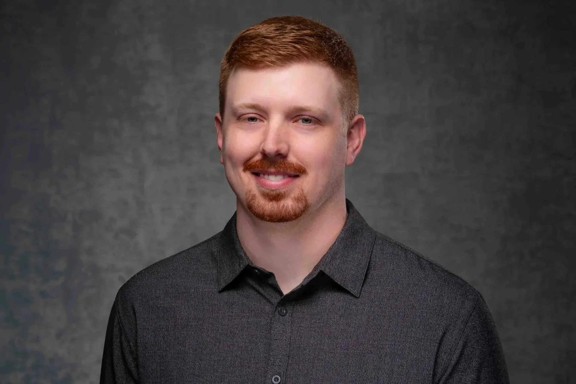 Headshot of a man with short red hair, a beard, and mustache, wearing a dark gray collared shirt, smiling slightly against a gray textured background.