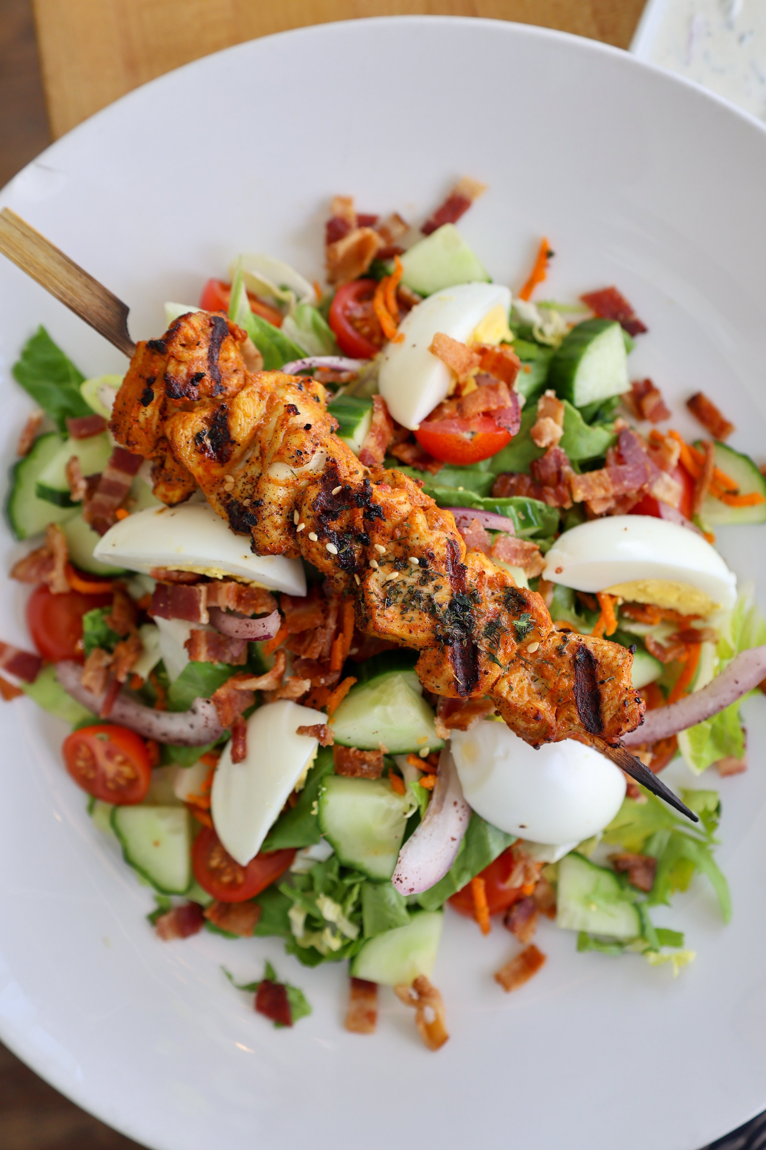 Mixed salad with cherry tomatoes, hard-boiled eggs, cucumber, carrots, bacon, and grilled chicken on a white plate.