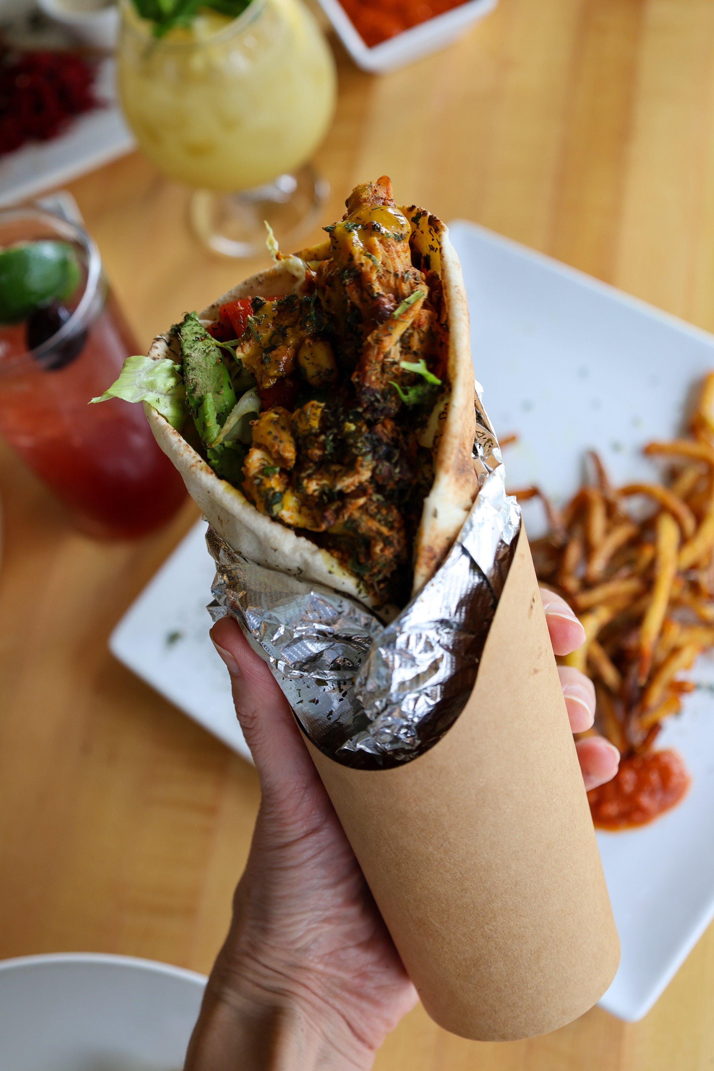A hand holding a wrapped burrito filled with grilled chicken, lettuce, and diced tomatoes. In the background, there is a glass of yellow-green beverage, a bowl of red salsa, and some fried noodles on a white plate on a wooden table.