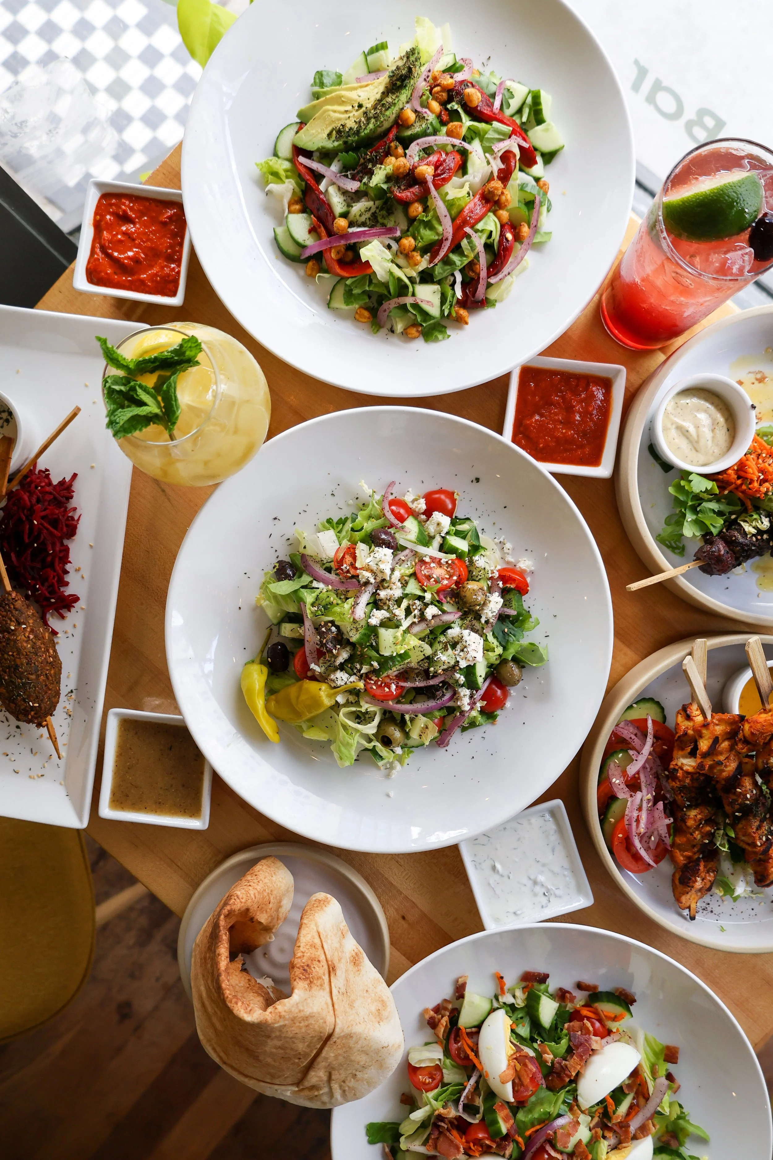 A variety of salads with fresh vegetables, olives, and cheese served on white plates, accompanied by small bowls of dressing and sauces, a glass of pink cocktail with lime and cherries, a yellow drink with mint, and a basket of bread rolls.