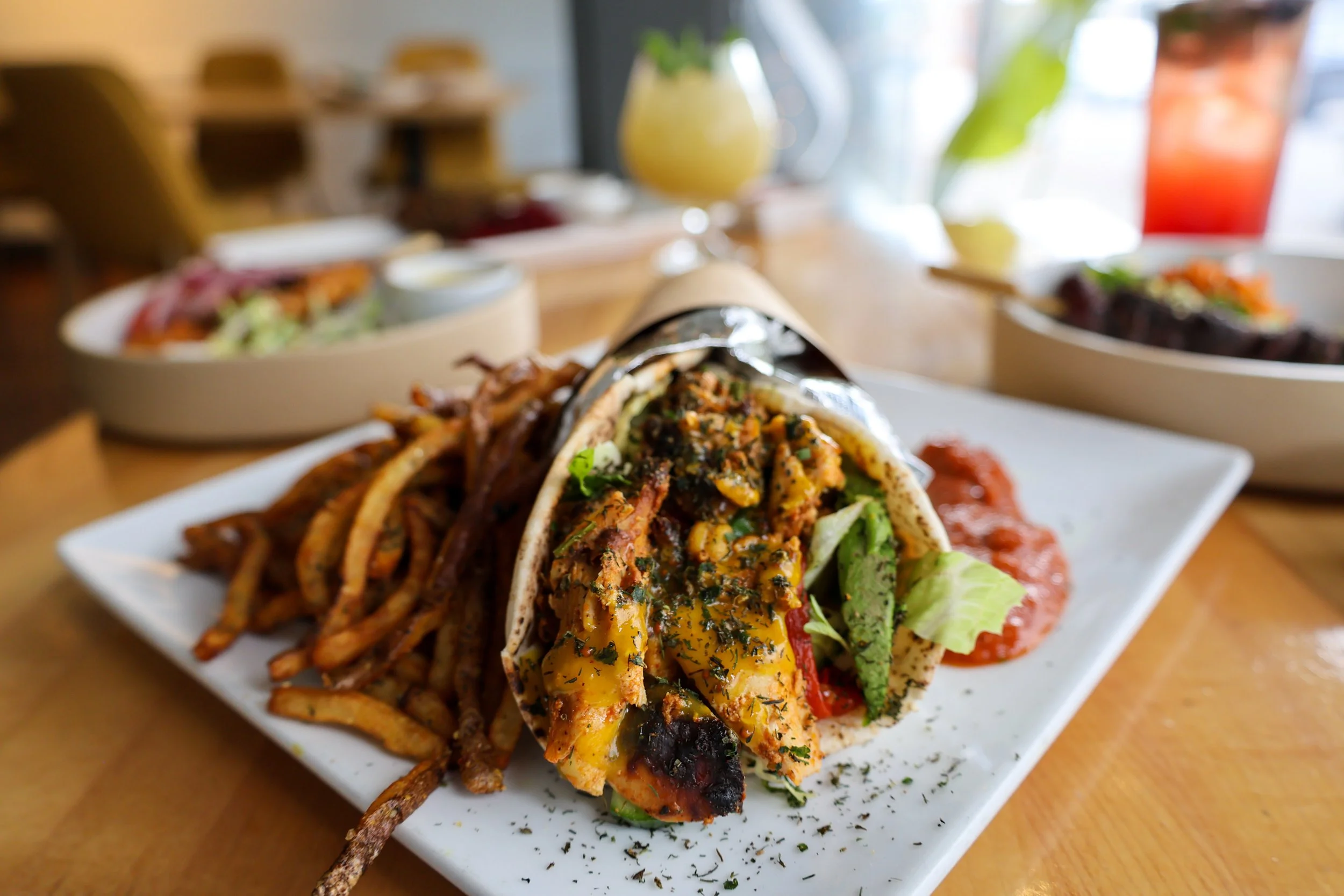 Close-up of a plate with a wrap sandwich filled with chicken, herbs, and vegetables, served alongside seasoned fries on a white rectangular plate, with additional dishes and drinks in the blurred background.