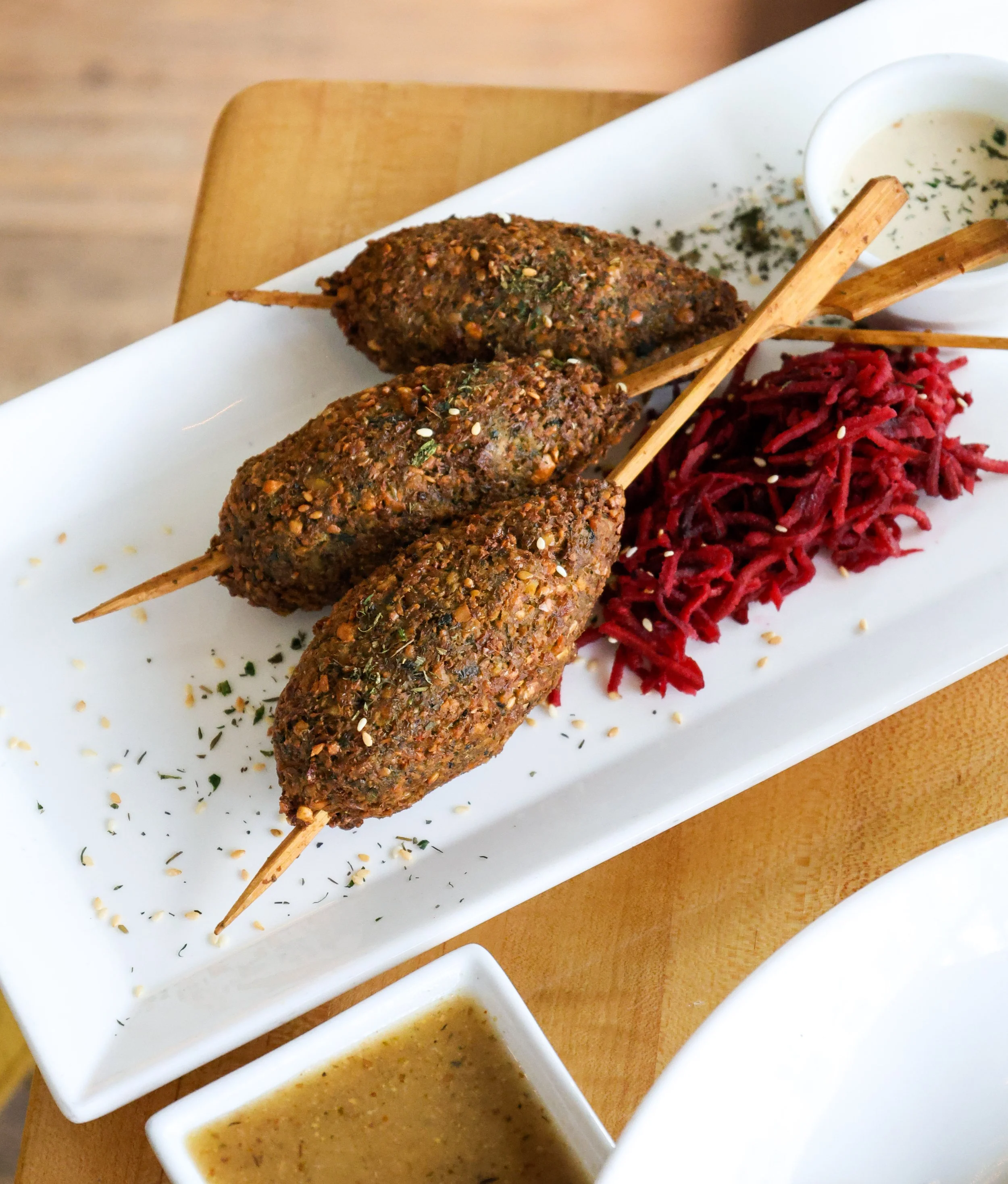 Three kebabs on skewers on a white rectangular plate, with a side of red shredded vegetables and a bowl of white sauce with herbs, and two additional dipping sauces in small white dishes.