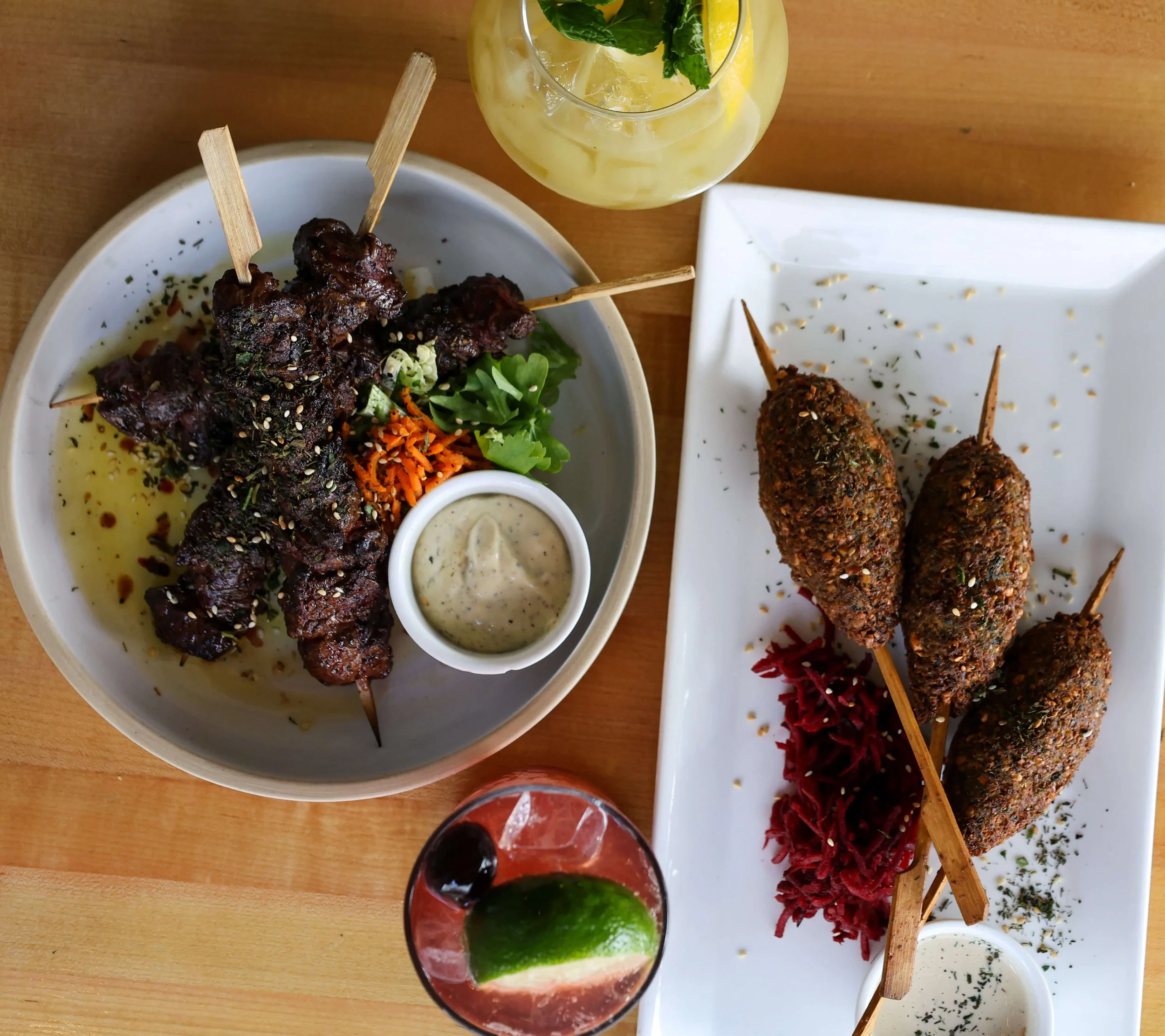 A variety of Mediterranean dishes on a wooden table, including skewered grilled meat, falafel, hummus with herbs, a bowl of creamy sauce, a mojito with mint and lemon, and a glass of dark soda.