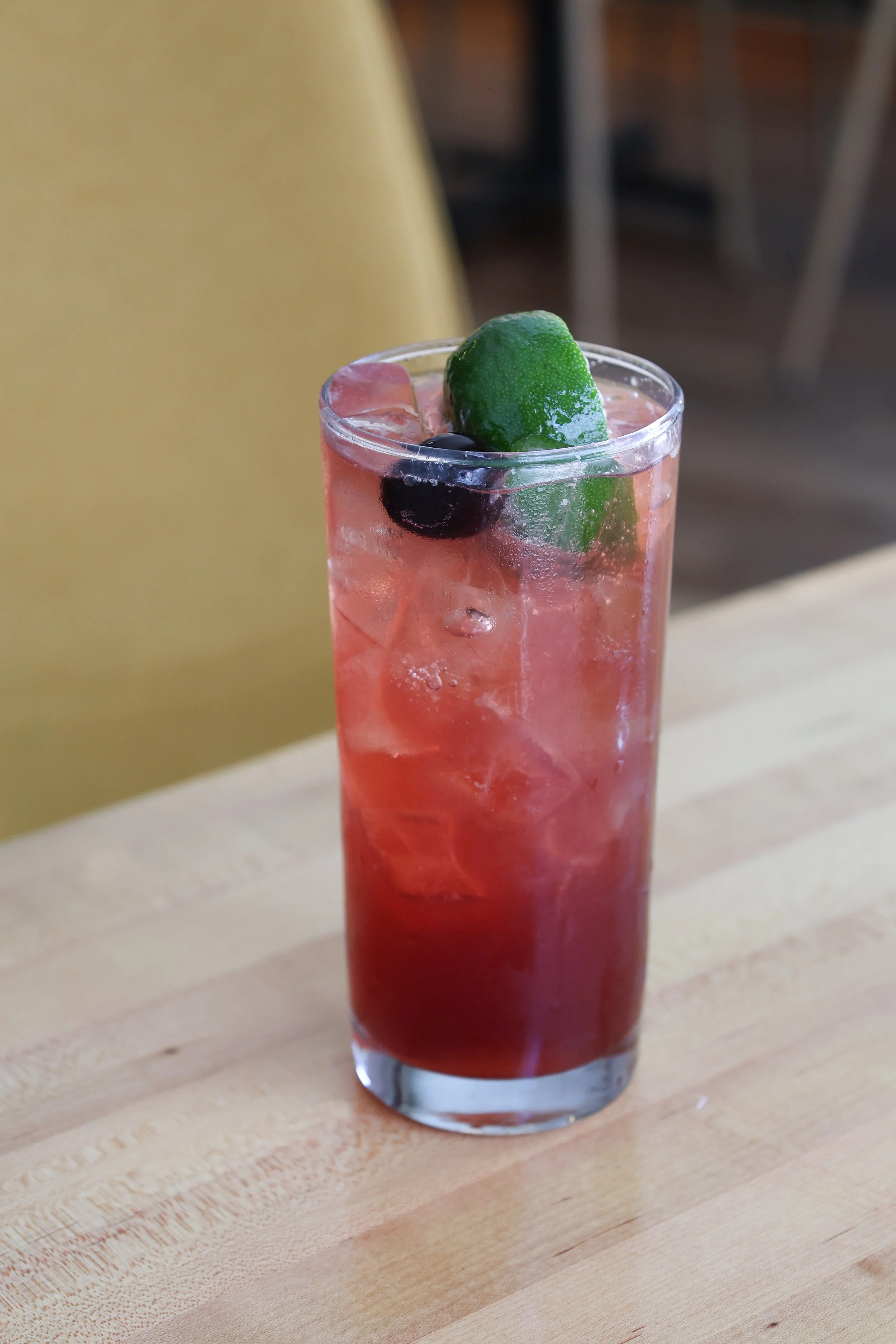 A tall clear glass filled with pink-colored beverage, ice cubes, a lime wedge, and a few dark berries on top, placed on a light wooden surface.