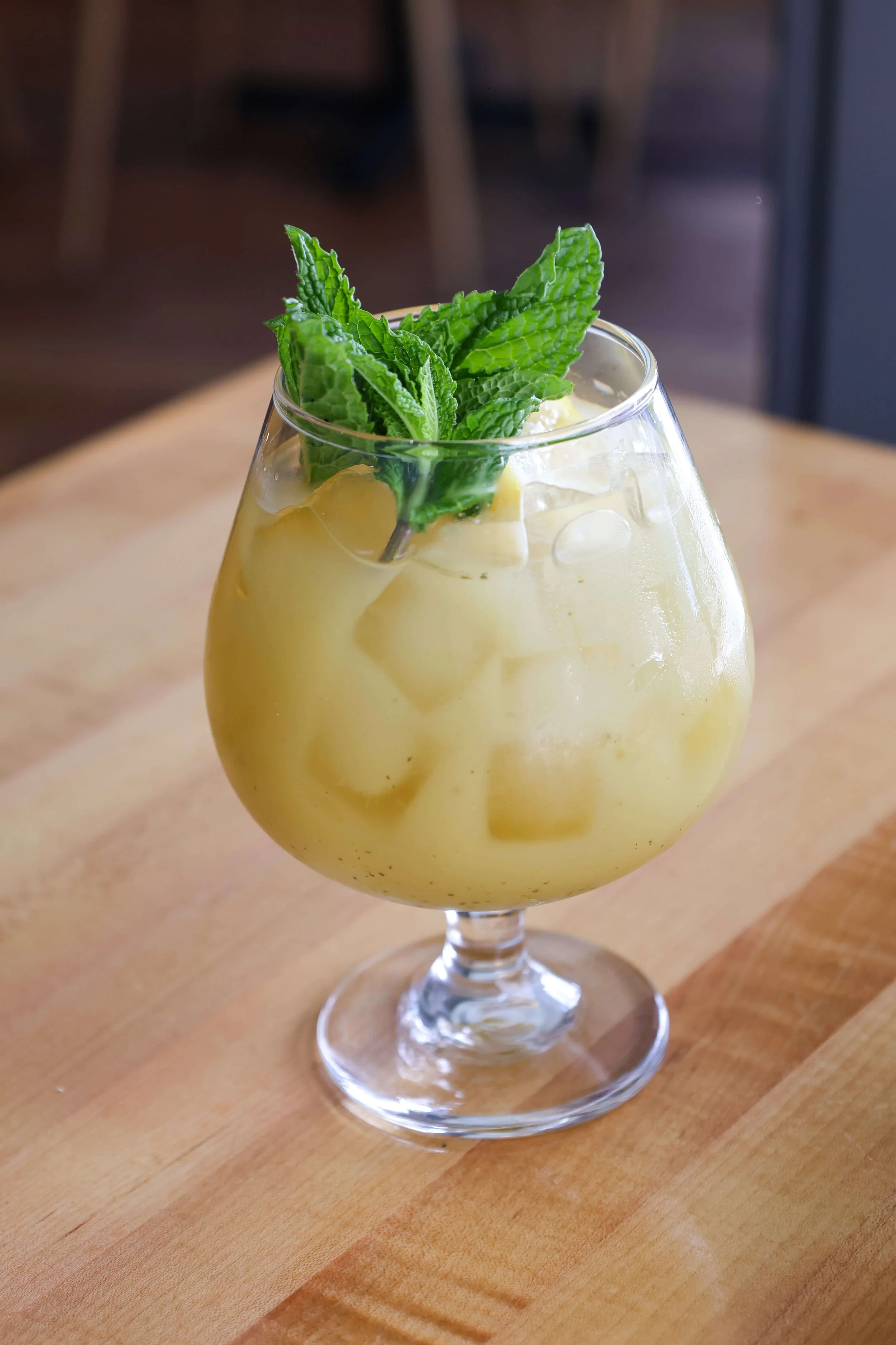 A glass of yellow-green beverage with ice and fresh mint leaves on top, placed on a wooden surface.
