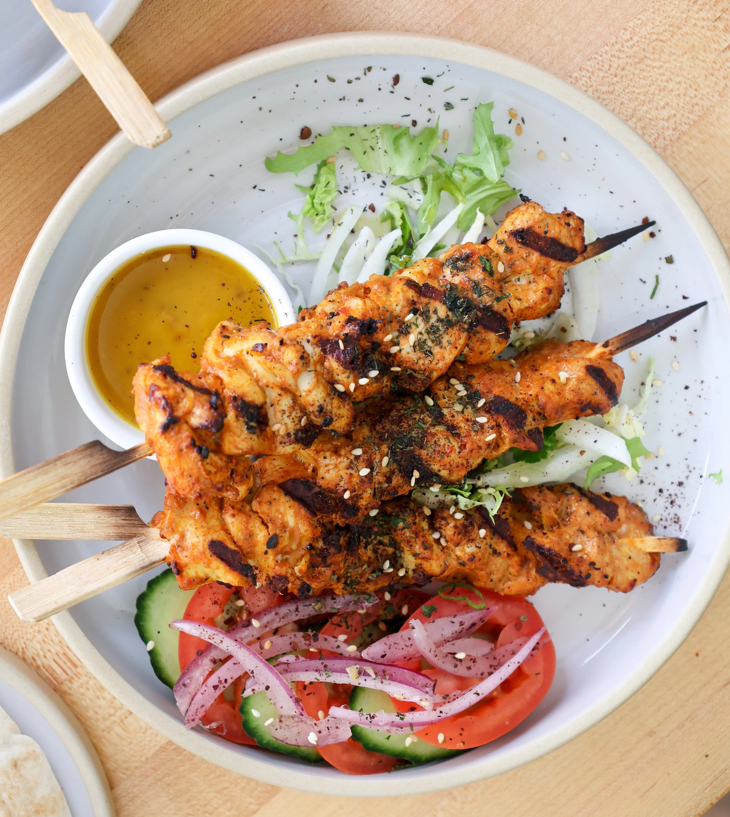 Close-up of a plate with grilled chicken skewer, fresh vegetable salad with sliced tomatoes, cucumbers, and onions, and a small bowl of yellow dipping sauce. The dish is garnished with sesame seeds and chopped herbs.
