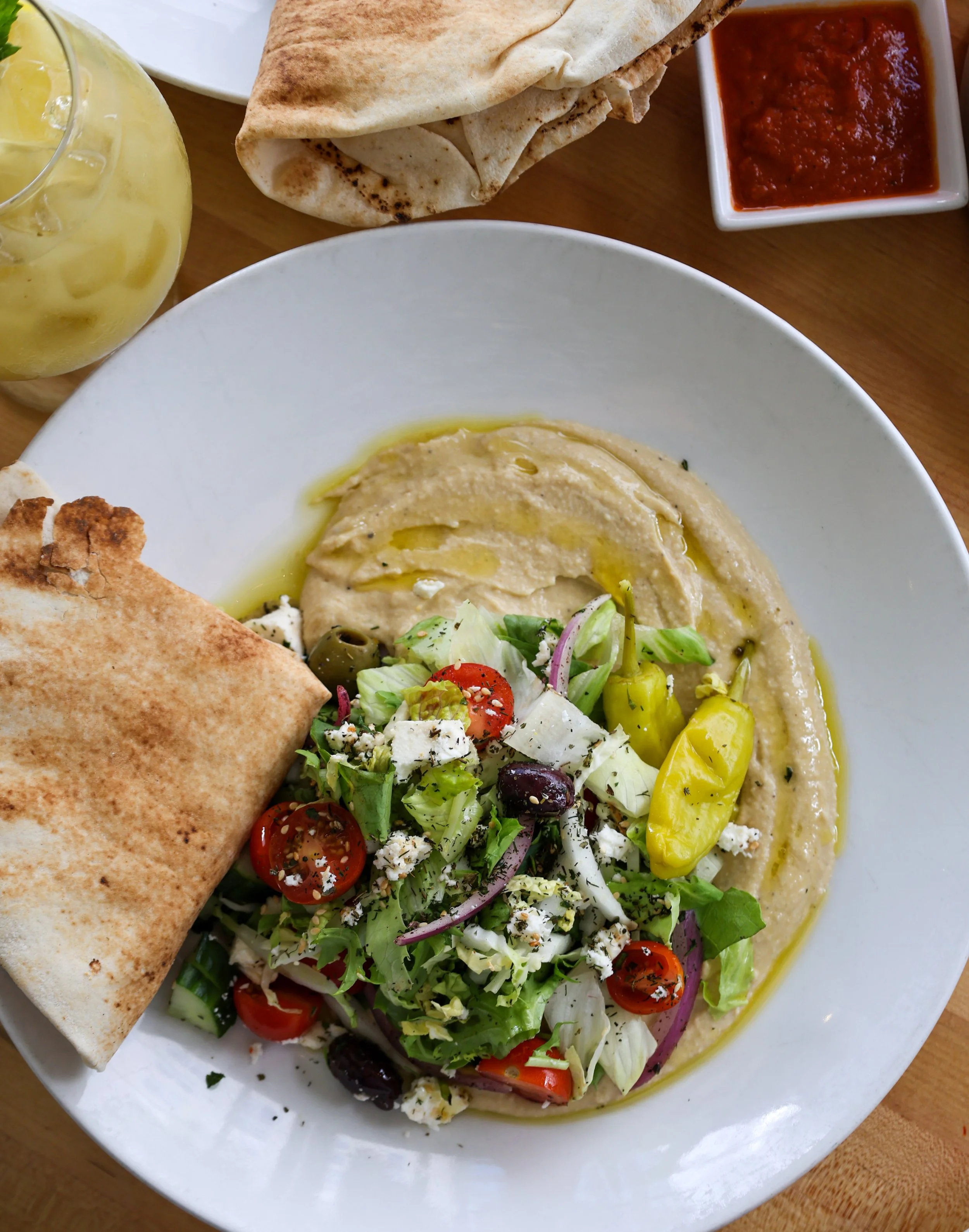 Plate of hummus topped with olive oil and spices, fresh salad with cherry tomatoes, cucumbers, red onions, olives, and feta cheese, along with a folded piece of flatbread, side of red sauce, and a glass of lemonade with ice.