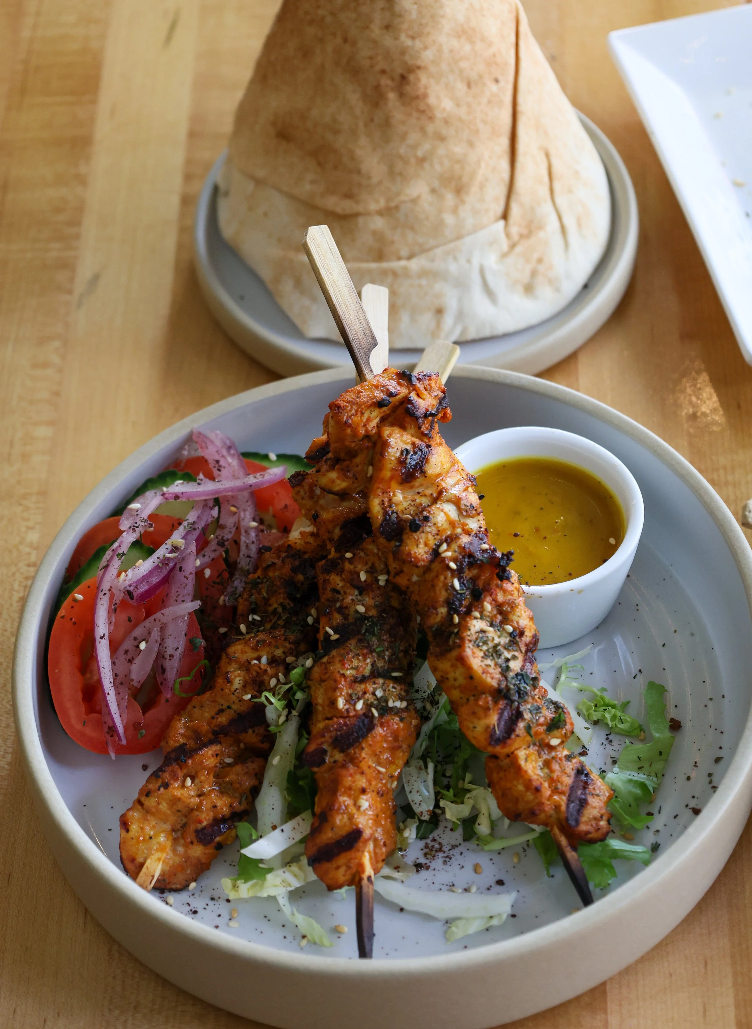 A plate of grilled chicken skewers with a side of fresh salad, served with a small bowl of yellow sauce. There is a bowl of pita bread in the background.