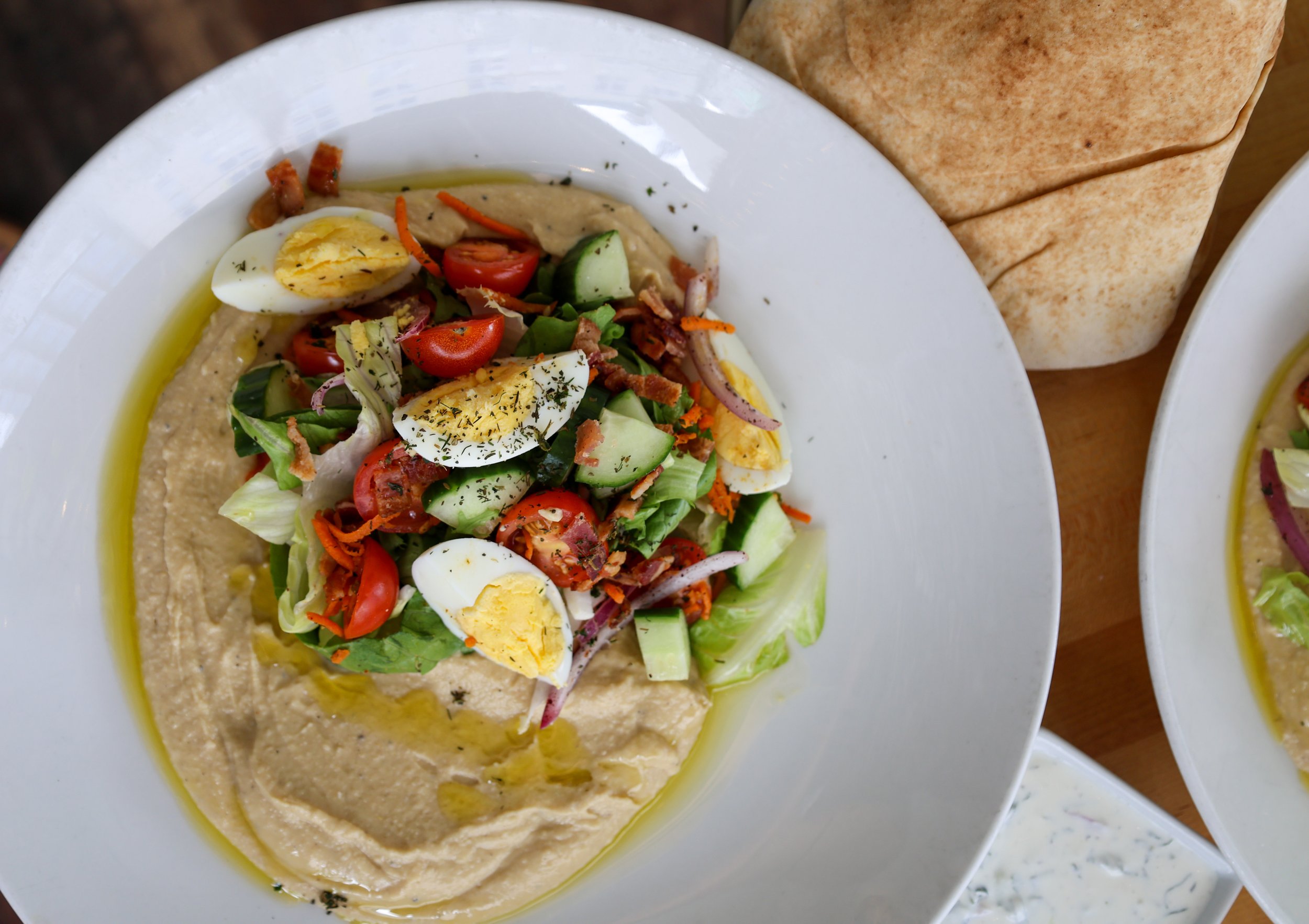 A plate of hummus topped with chopped vegetables, cherry tomatoes, sliced boiled eggs, and herbs, served with a side of flatbread.