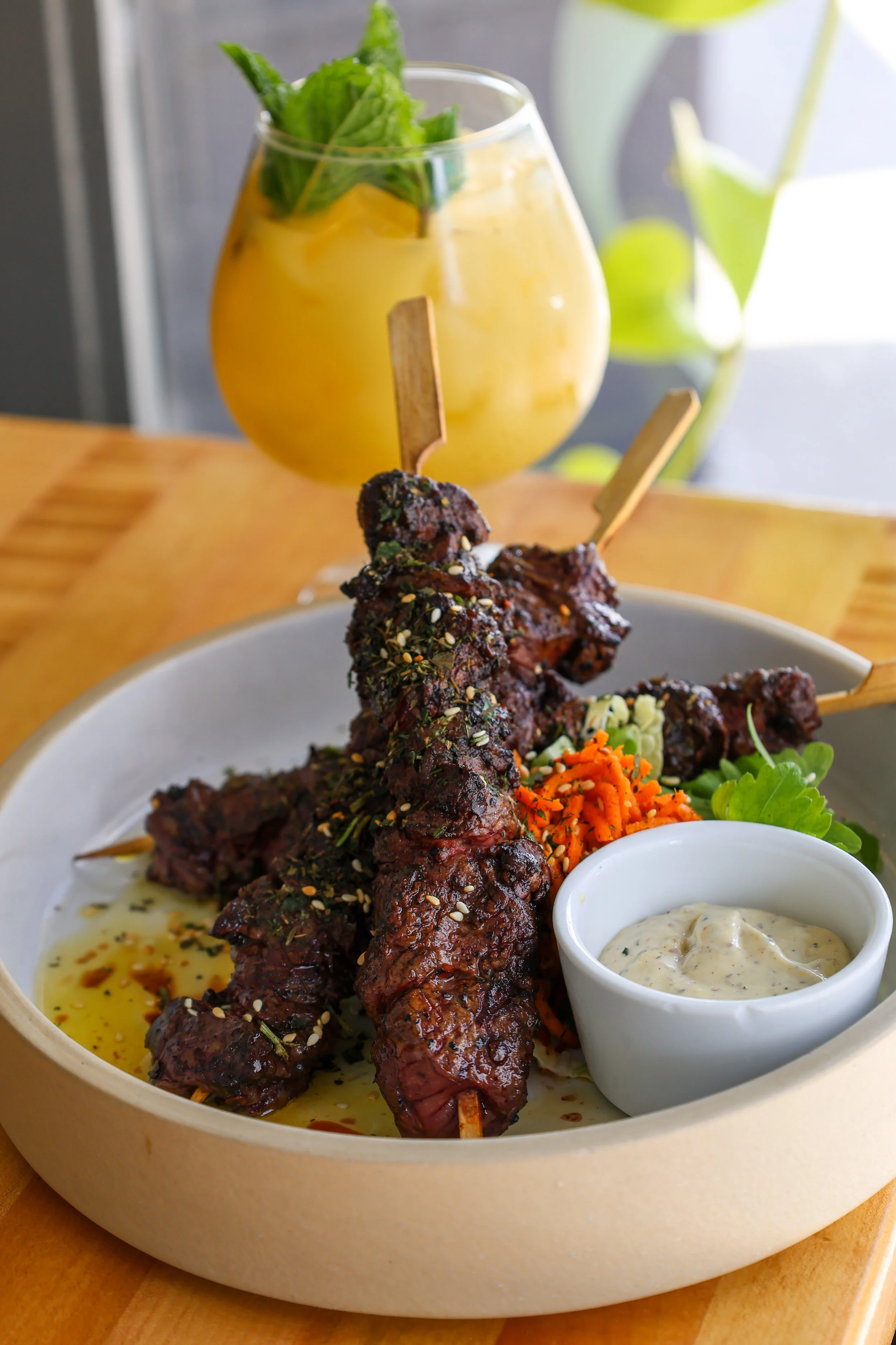 Plate of grilled beef skewers topped with sesame seeds, served with a side of sauce, shredded carrots, and greens. A glass of yellow beverage with mint leaves is in the background.