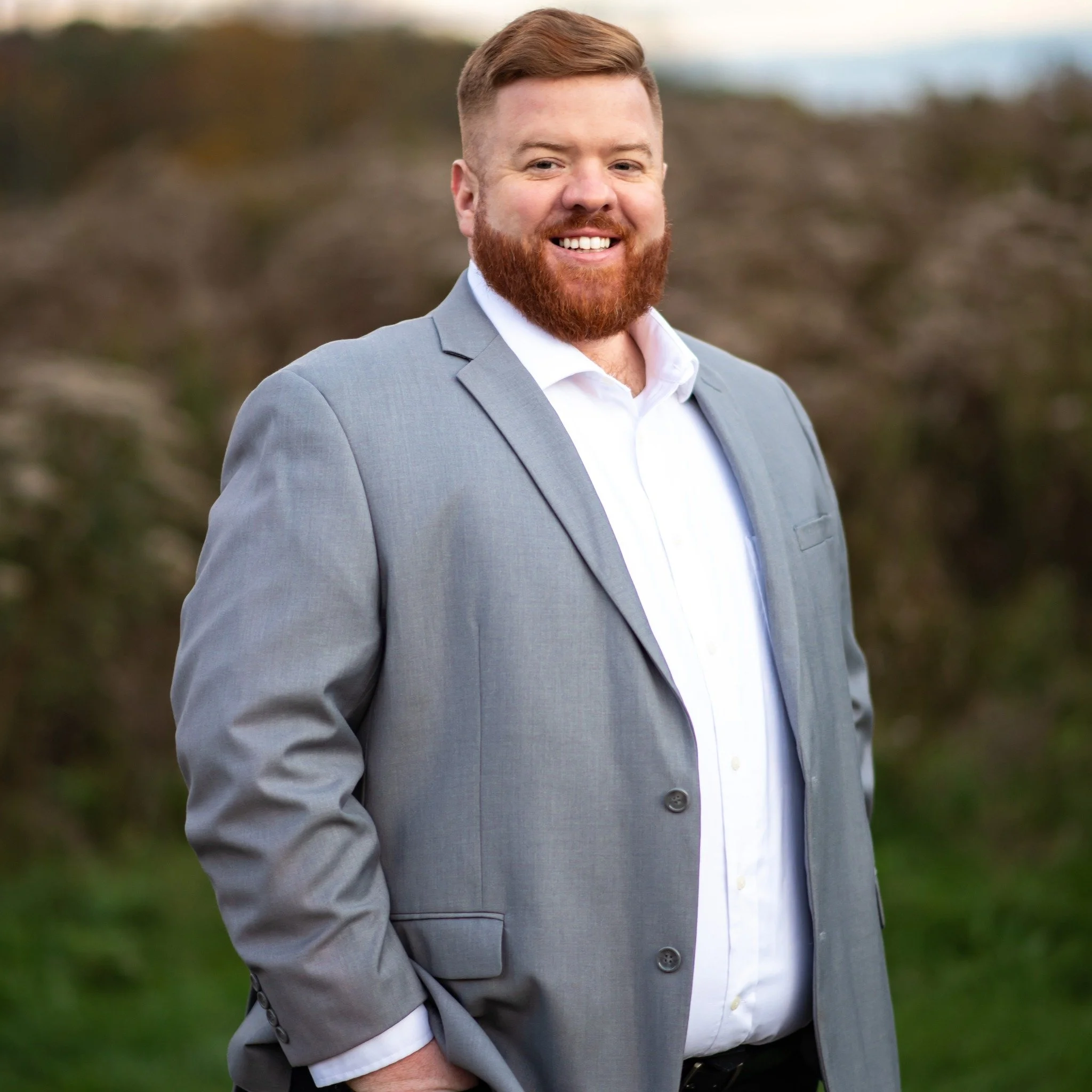 A smiling man with red hair, a beard, wearing a light gray suit and white shirt, standing outdoors with blurred nature background.
