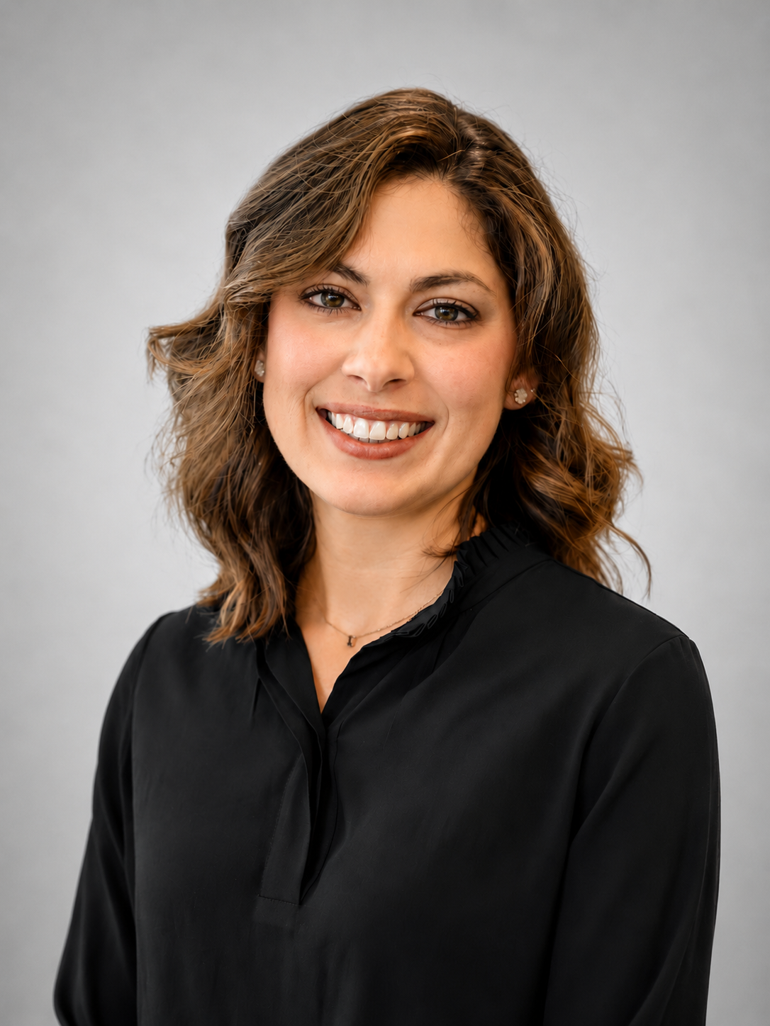 A woman with short, brunette hair wearing a black top and smiling, against a soft gray studio background.