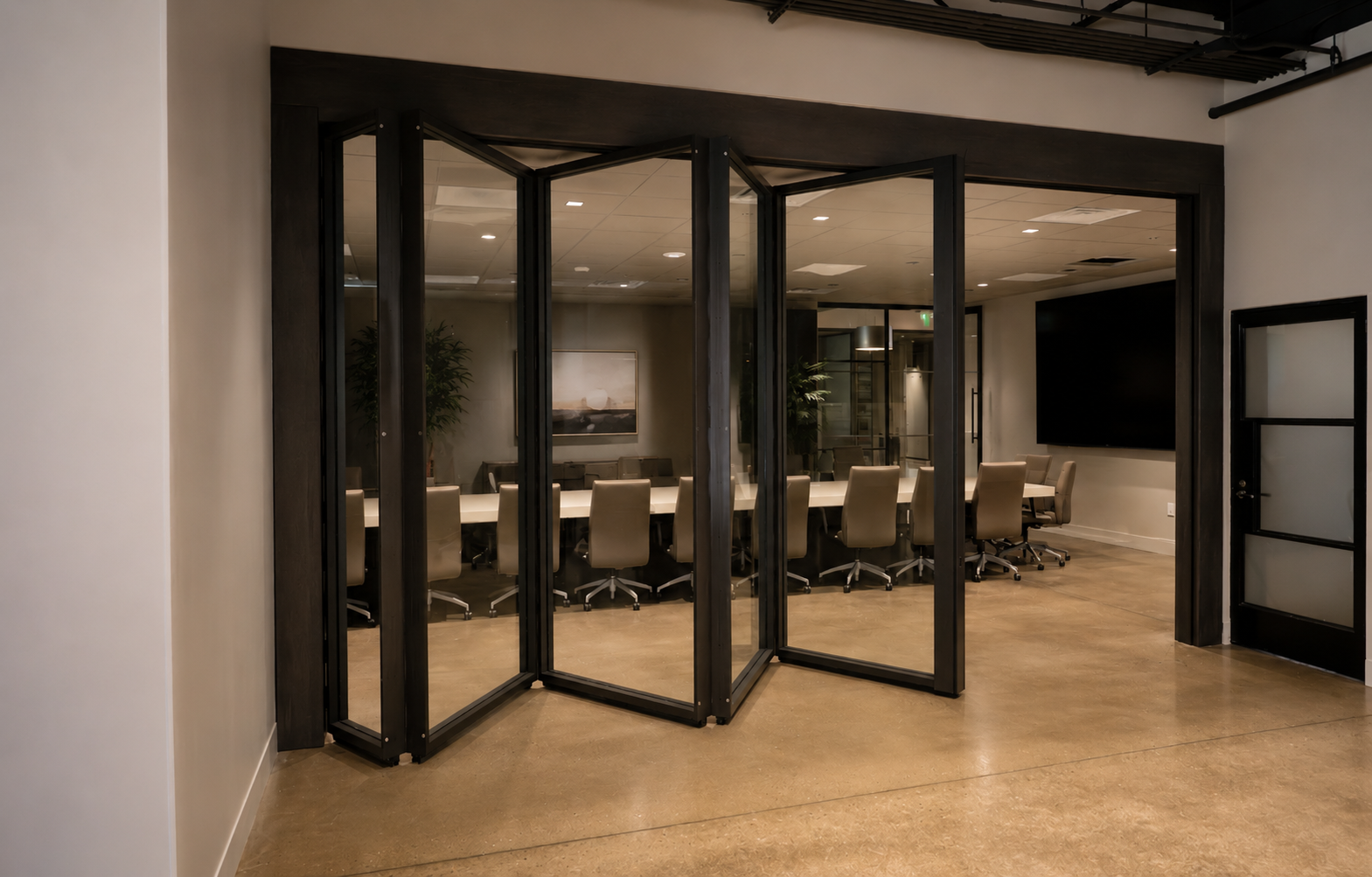 Conference room with beige chairs around a white table, viewed through black-folding glass doors in an office setting.