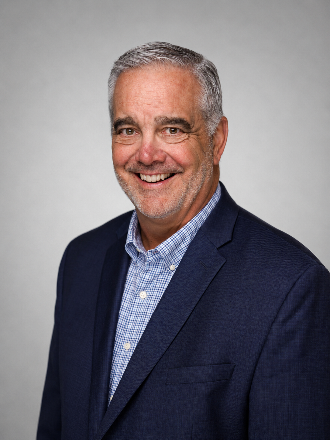 Portrait of a man with slightly grey hair wearing a blue and white shirt and a dark blue blazer, smiling against a light gray background.