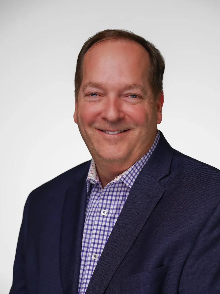 A man with light skin, short brown hair, and blue eyes smiling, wearing a navy blazer over a purple and white checkered shirt, against a plain white background.