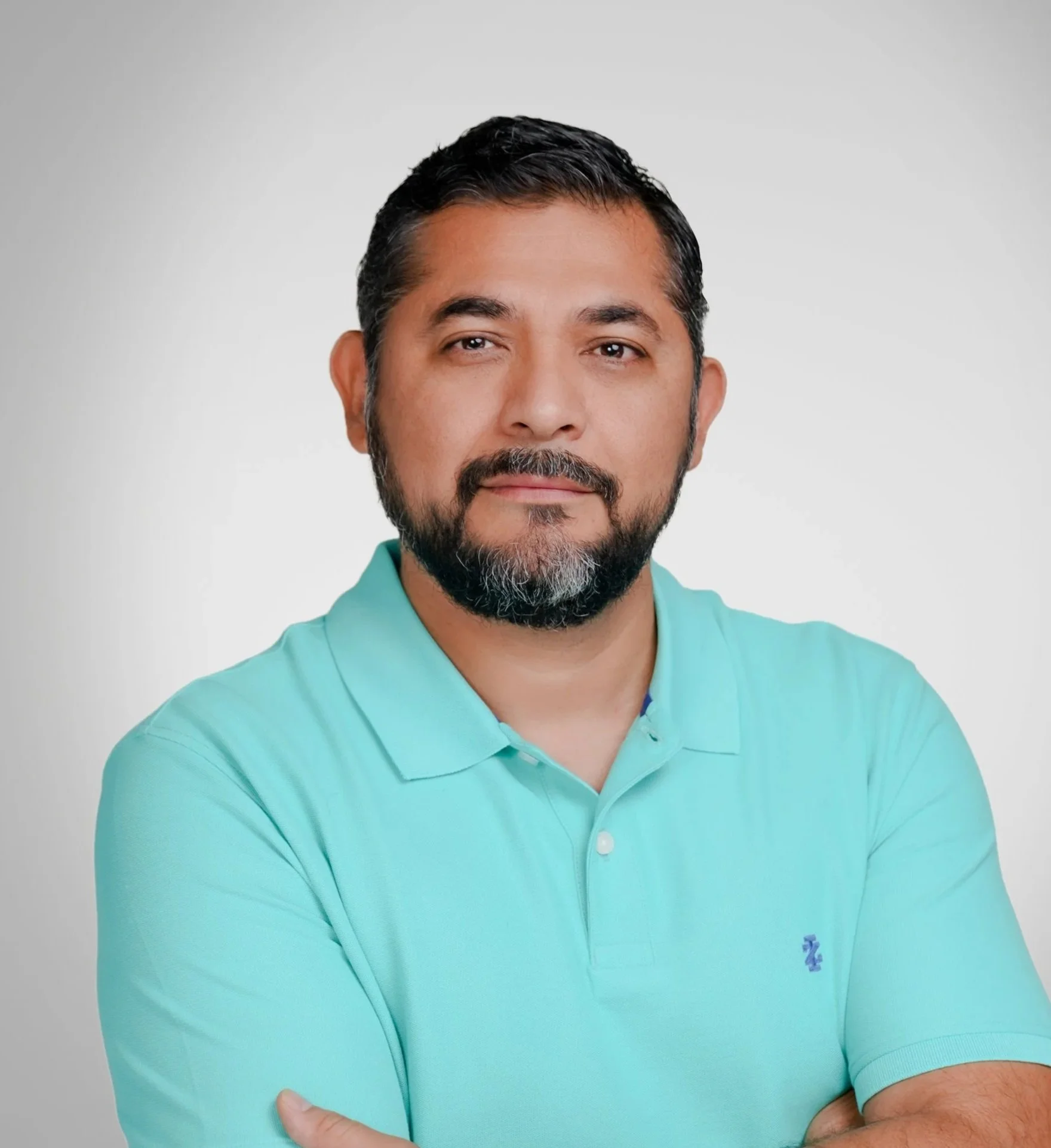 Portrait of a man with dark hair, beard, and mustache, wearing a light teal polo shirt with a small logo, standing against a plain light gray background.