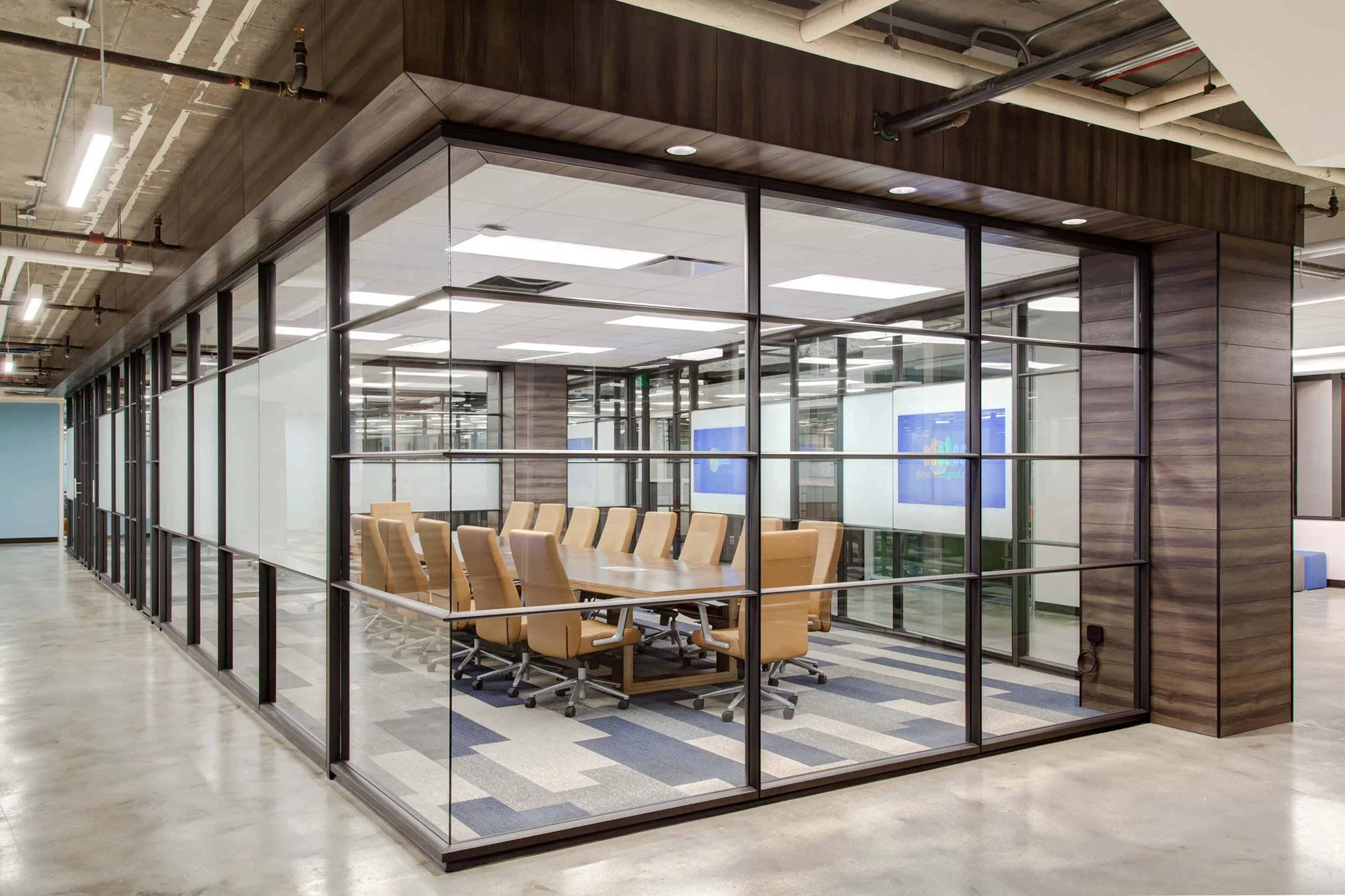 A glass-walled conference room in an office building with a long table and tan chairs inside. The room has a wooden trim at the top and visible ceiling pipes and lighting fixtures.