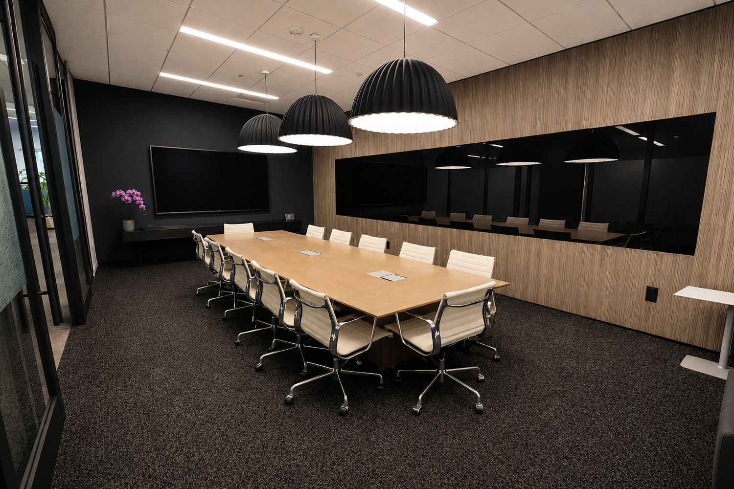 Empty conference room with a large wooden table, white chairs, black pendant lights, black wall with a large screen, and a potted purple orchid.