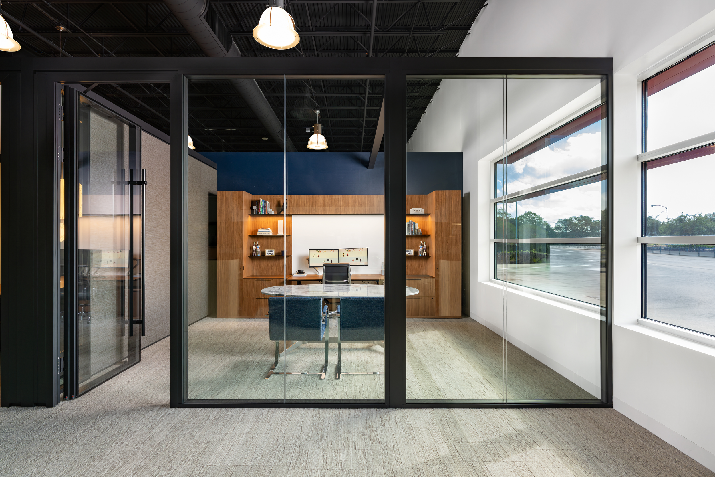 Office workspace seen through a glass wall with black framing, featuring a wooden desk with two computer monitors, a chair, and shelves with books, illuminated by large windows and ceiling lights.