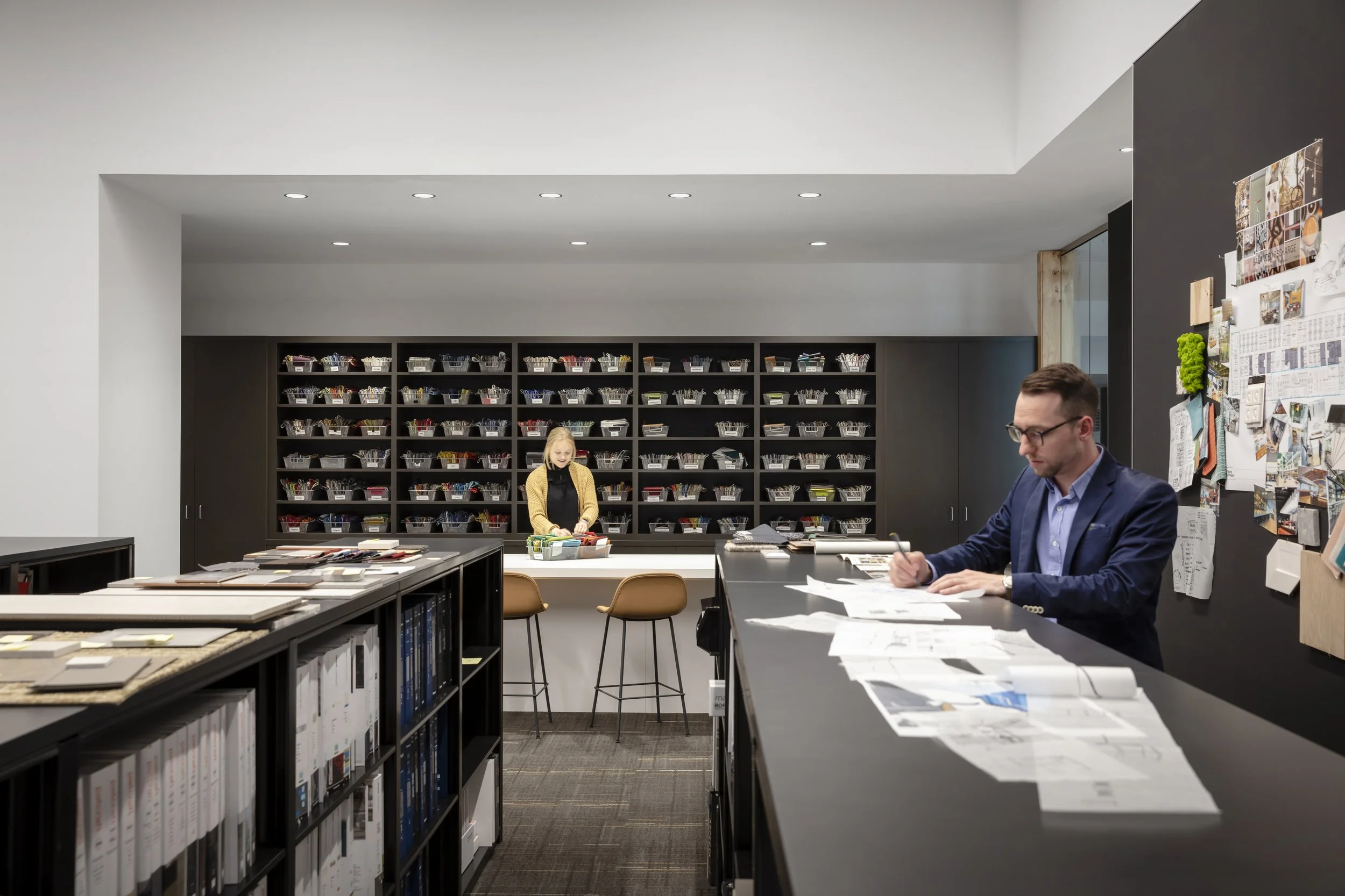A modern office or design studio with two people working at a large table. There are shelves with organized compartments containing papers and supplies, a black wall with various pictures and notes pinned on it, and a white wall with ceiling lights.