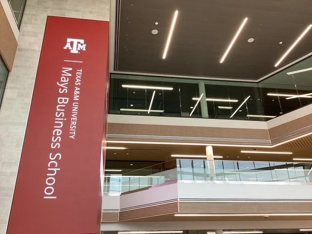 Interior view of Texas A&M University's Mays Business School, featuring a red vertical sign with the university's logo and name, multiple stories with modern design elements, and glass railings.