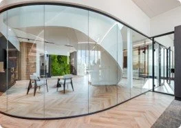 Modern living space with curved glass walls, hardwood flooring, and minimalist furniture, overlooking a green outdoor area.