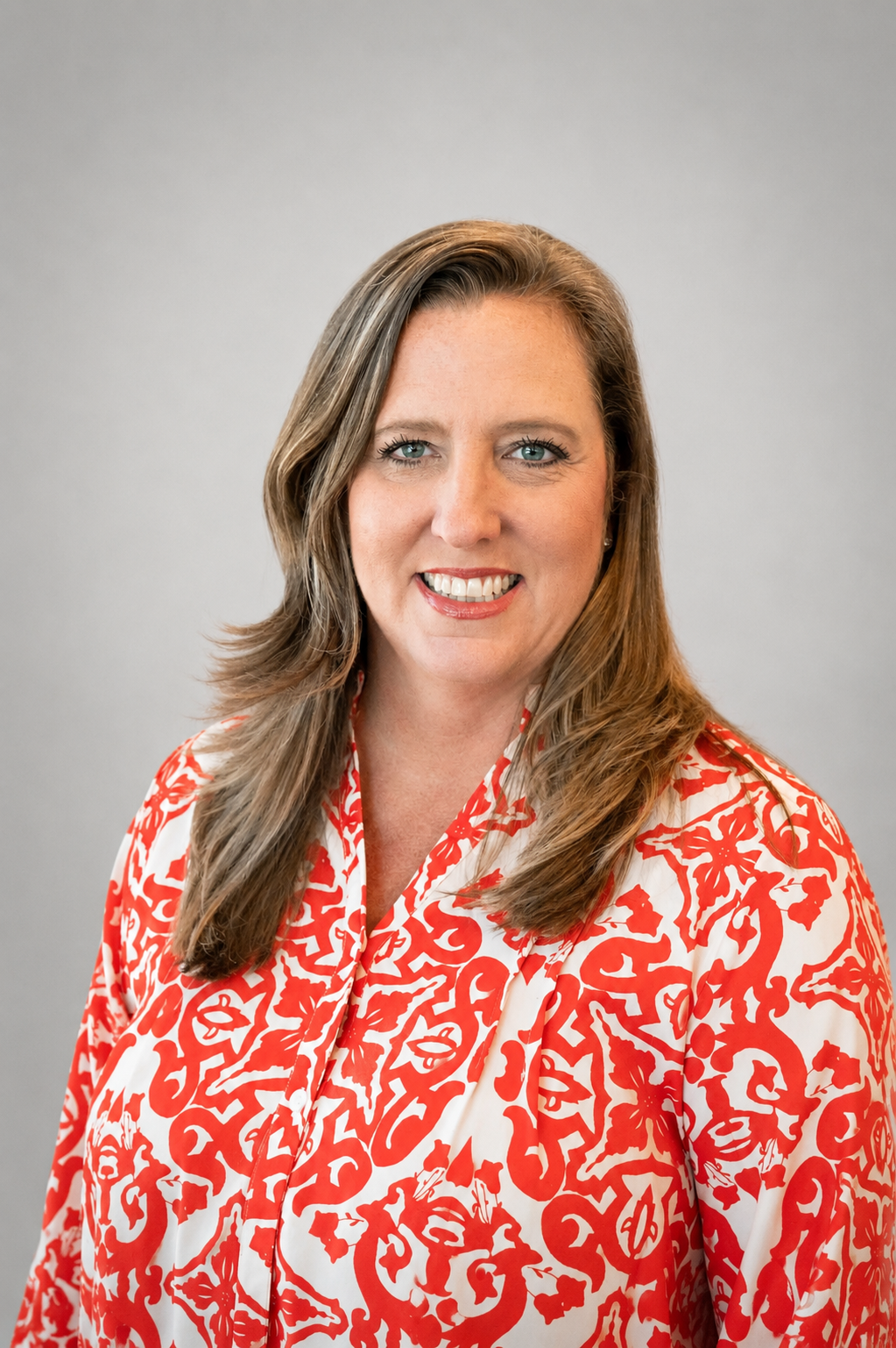 A woman with light brown hair and blue eyes smiling, wearing a white blouse with a red floral pattern, against a plain grey background.