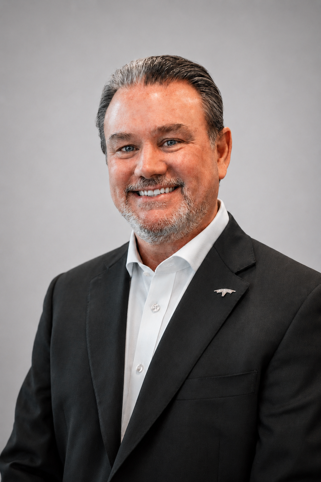 Headshot of a middle-aged man with gray hair and beard, wearing a black suit jacket with a small pin, white shirt, smiling against a plain gray background