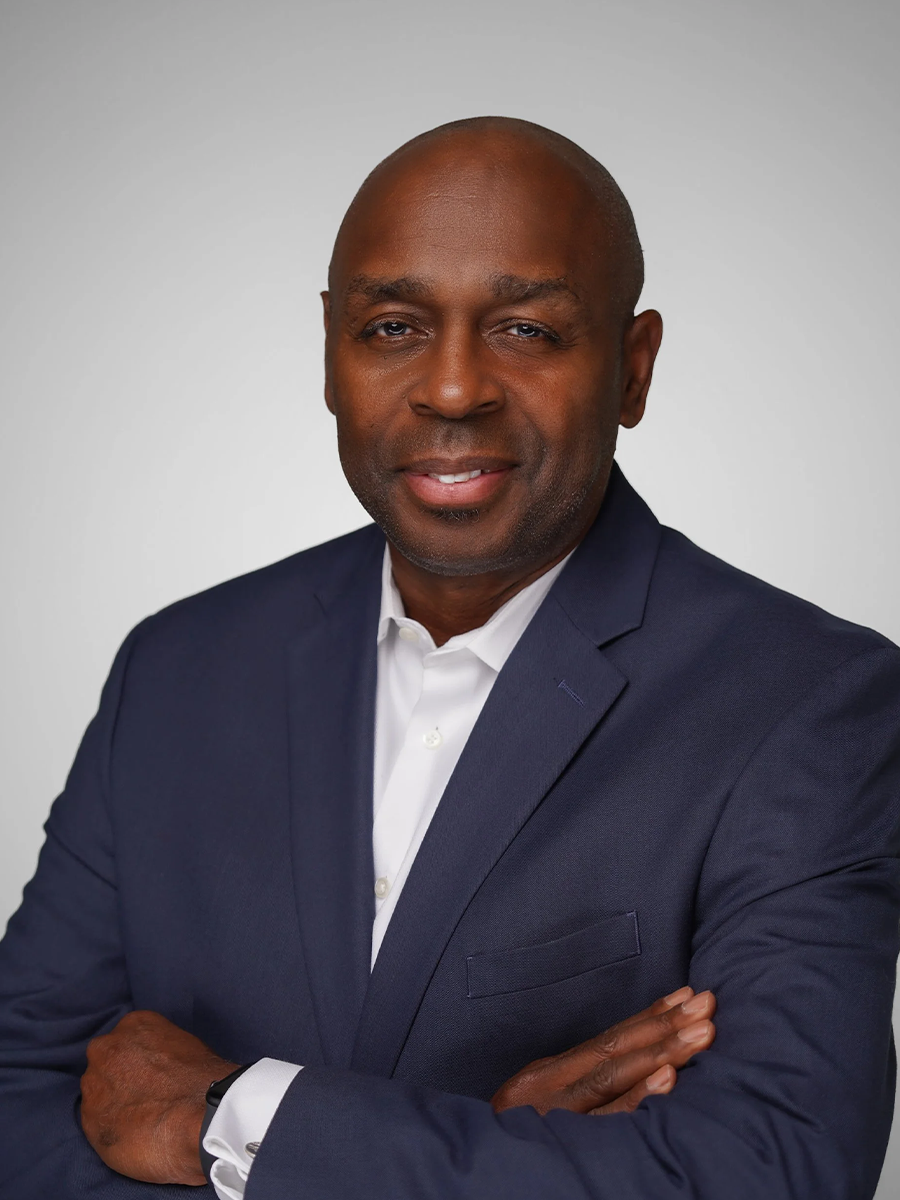 Headshot of a confident African American man in a navy suit with a white shirt, standing with arms crossed against a plain background.