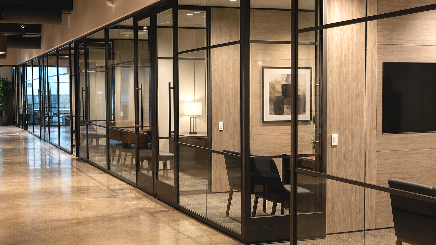 Modern office interior with glass-walled meeting rooms, wooden wall panels, black chairs, a framed art piece, and a flat-screen TV mounted on the wall.