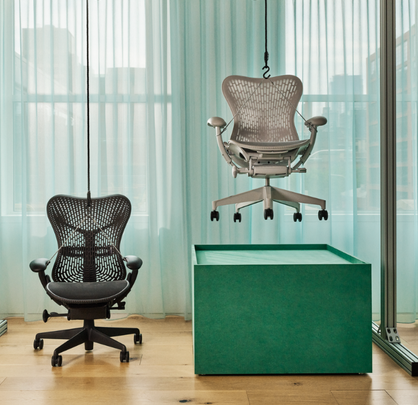Two modern ergonomic office chairs, one black and one white, in a bright office space with sheer curtains, wooden floor, and a green display platform.
