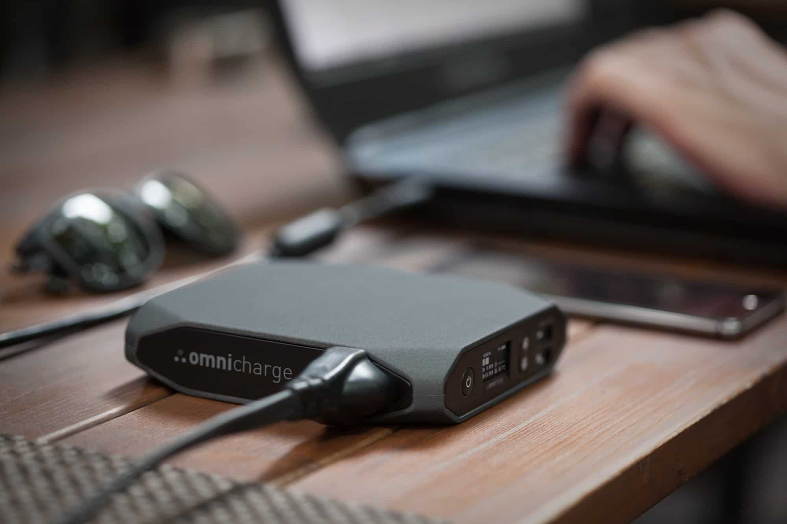 Portable black Omnicharge power bank connected to a charging cable on a wooden desk with headphones, a smartphone, and a laptop in the background.