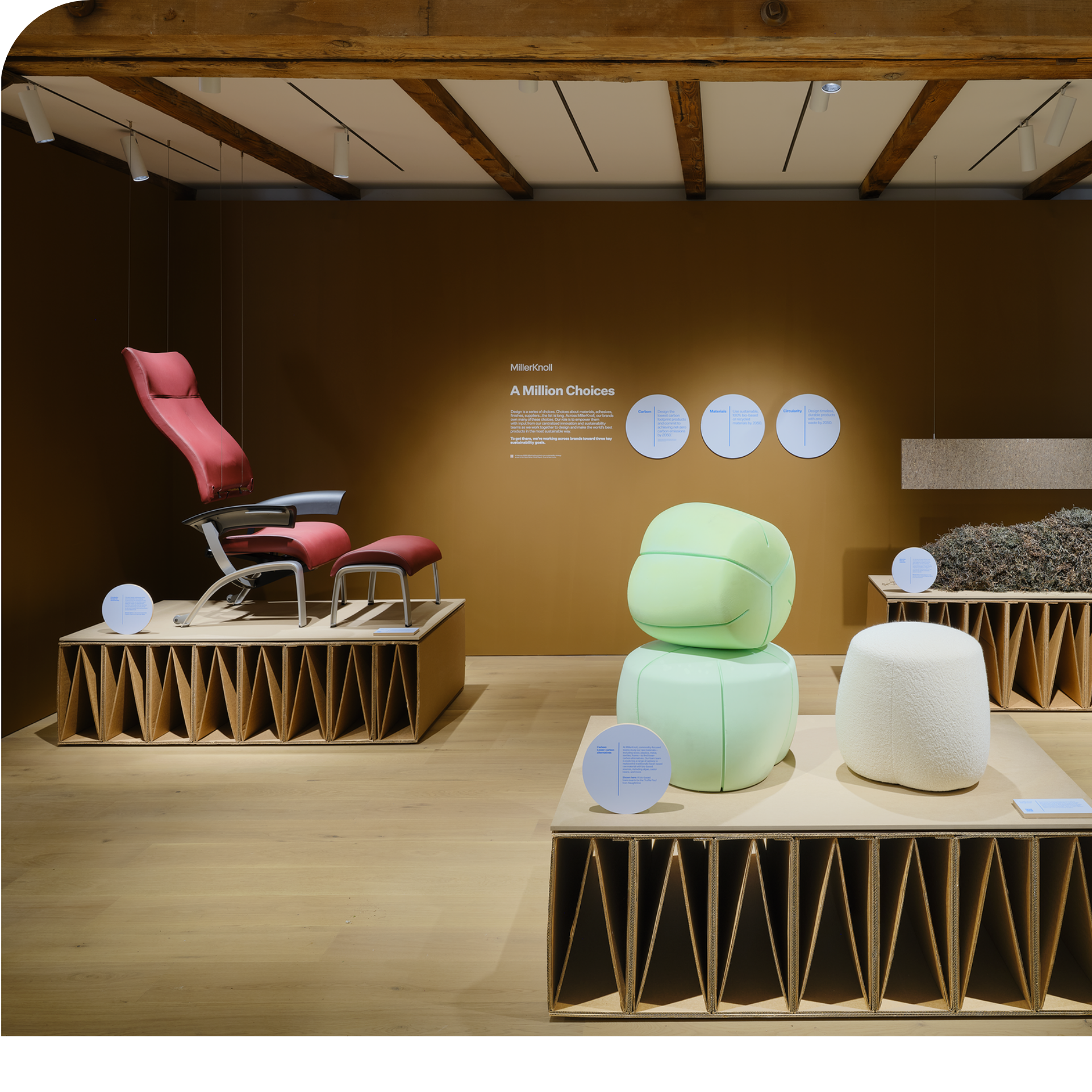 An exhibit displaying modern furniture, including a pink and grey reclining chair, a light green sculpture or ottoman, and a cream pouf, all set on wooden platforms against a brown wall with informational text.