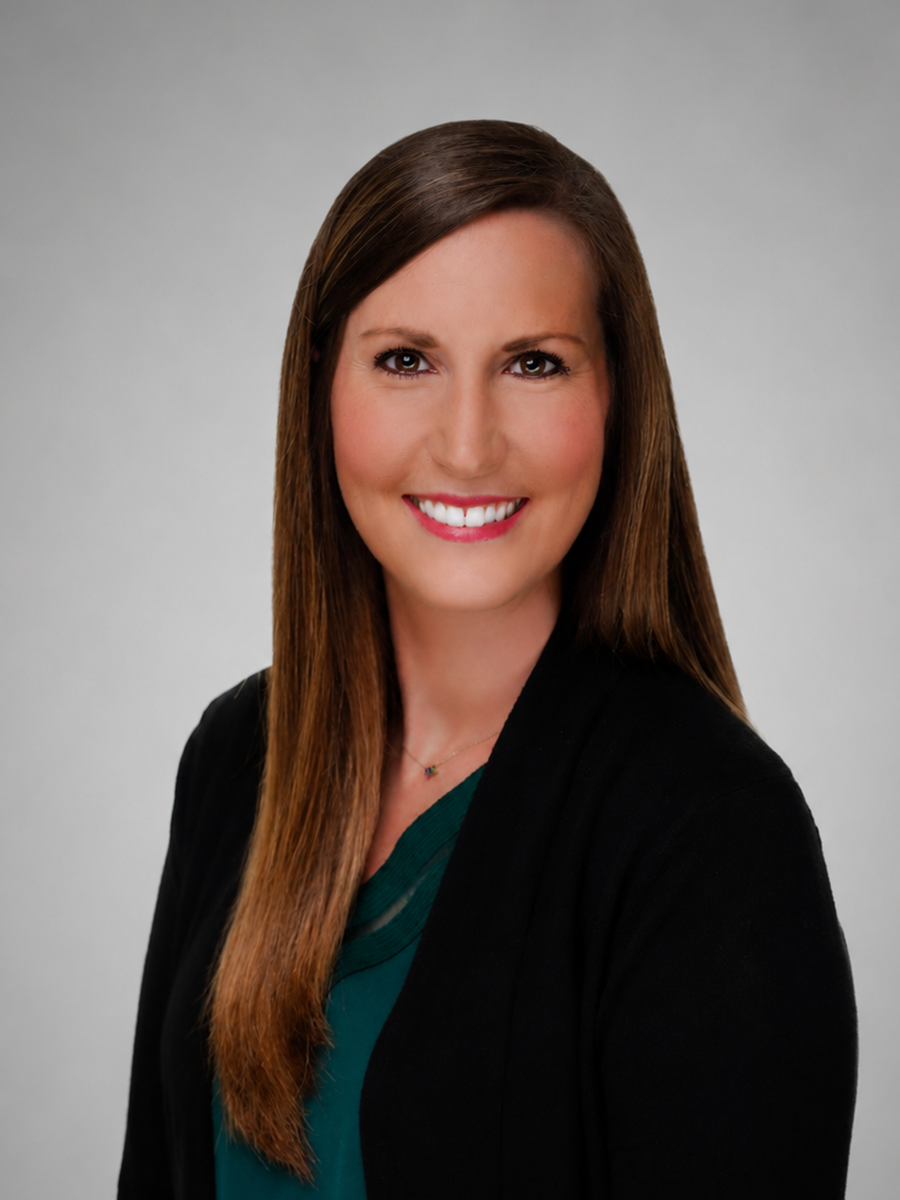 Professional headshot of a woman with long brown hair, smiling, wearing a dark blazer over a teal top, against a gray background.