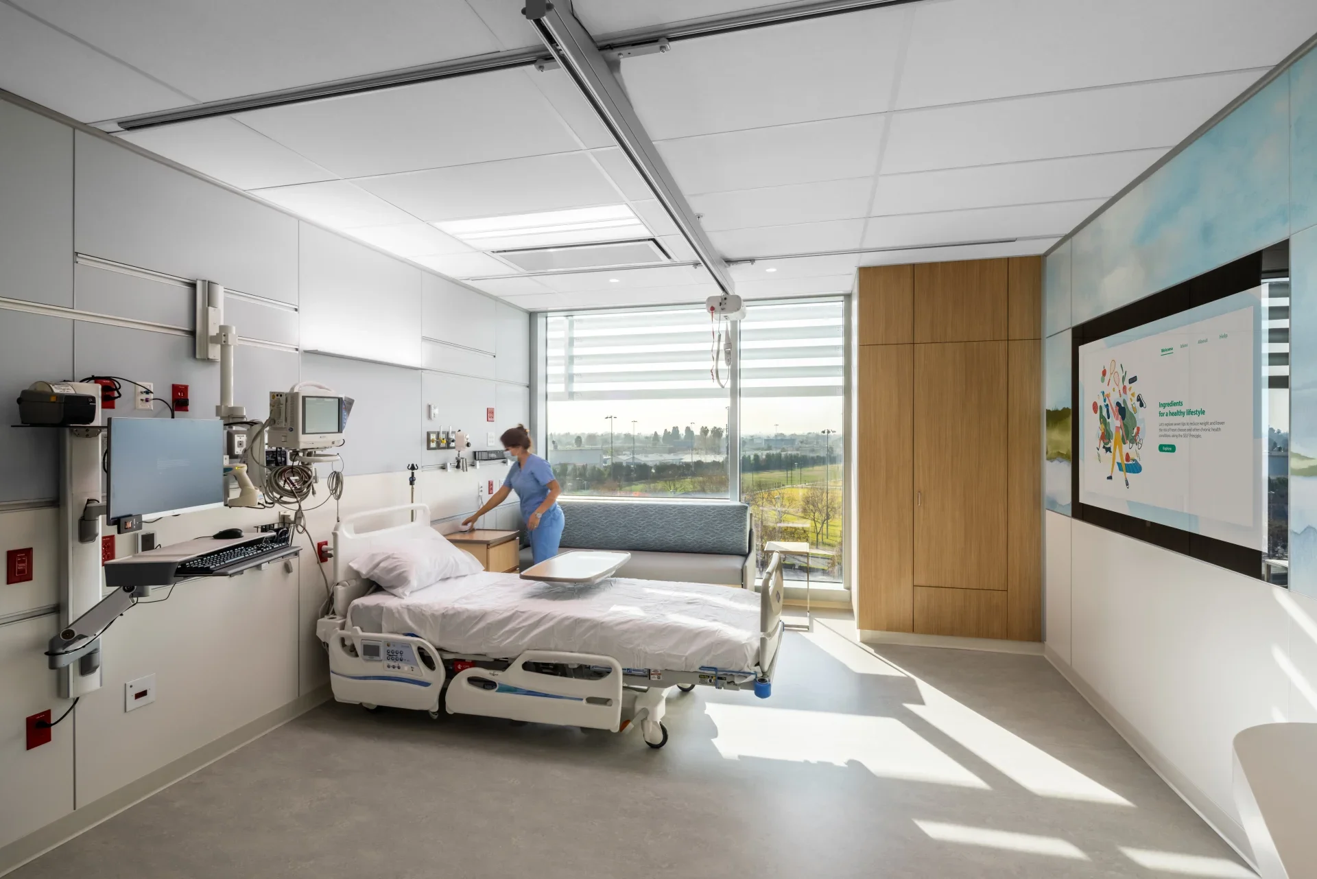 A hospital room with a bed, medical equipment, and a healthcare worker in scrubs cleaning the bed. Large windows with blinds letting in sunlight, and a flat screen TV on the wall.