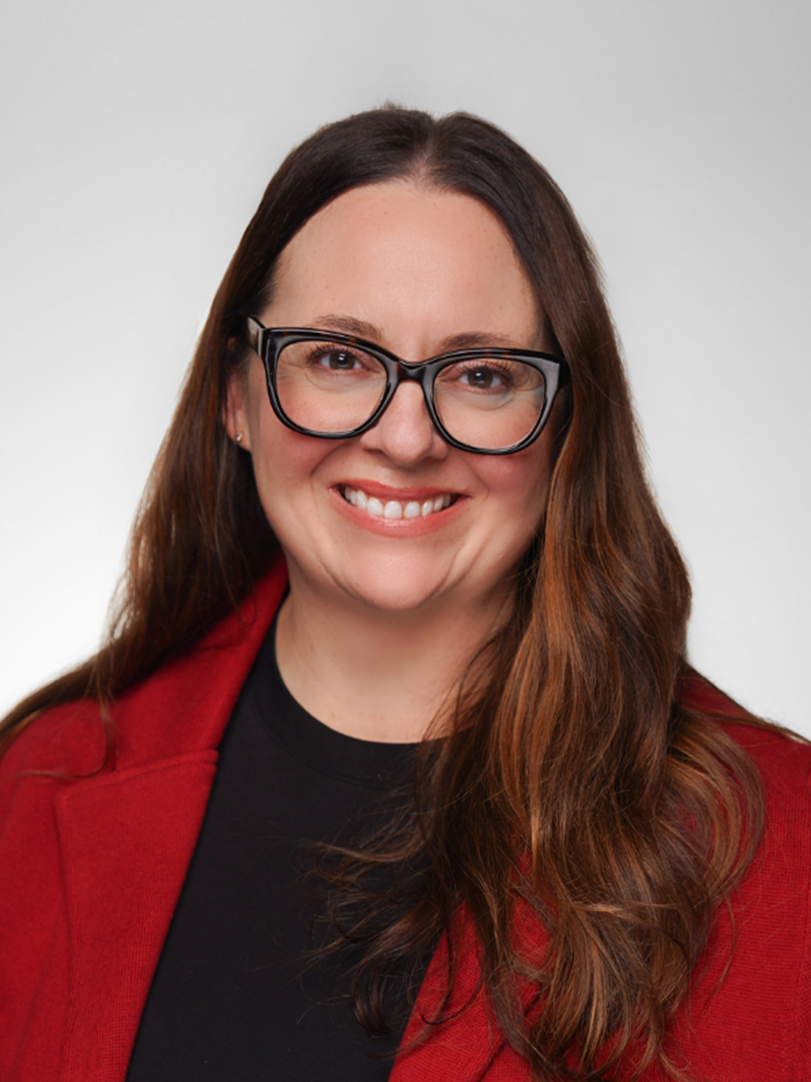 A woman with long brown hair wearing glasses, a black top, and a red blazer, smiling at the camera against a plain white background.