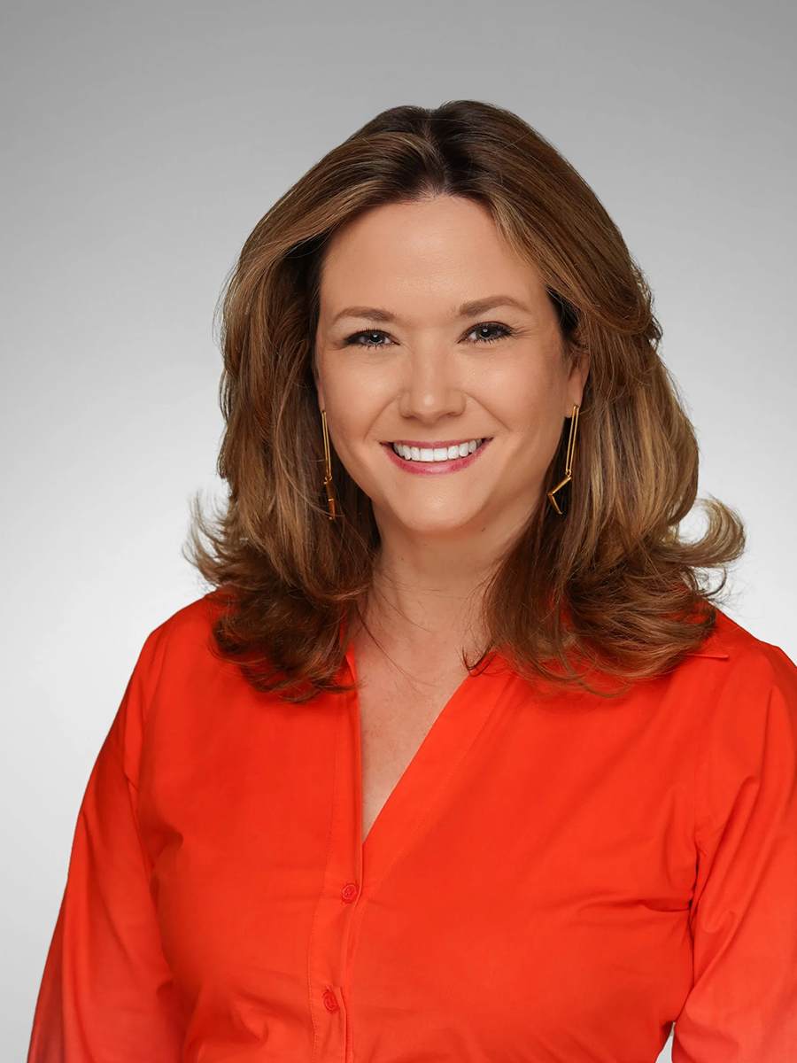 A woman with shoulder-length wavy brown hair, smiling, wearing a bright red blouse and gold earrings, against a neutral gray background.
