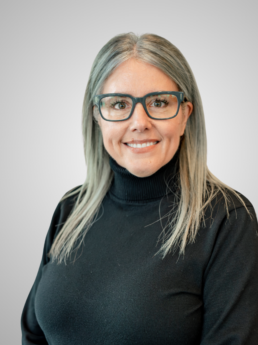 Portrait of a woman with shoulder-length gray hair, wearing glasses and a black turtleneck, smiling against a neutral background.
