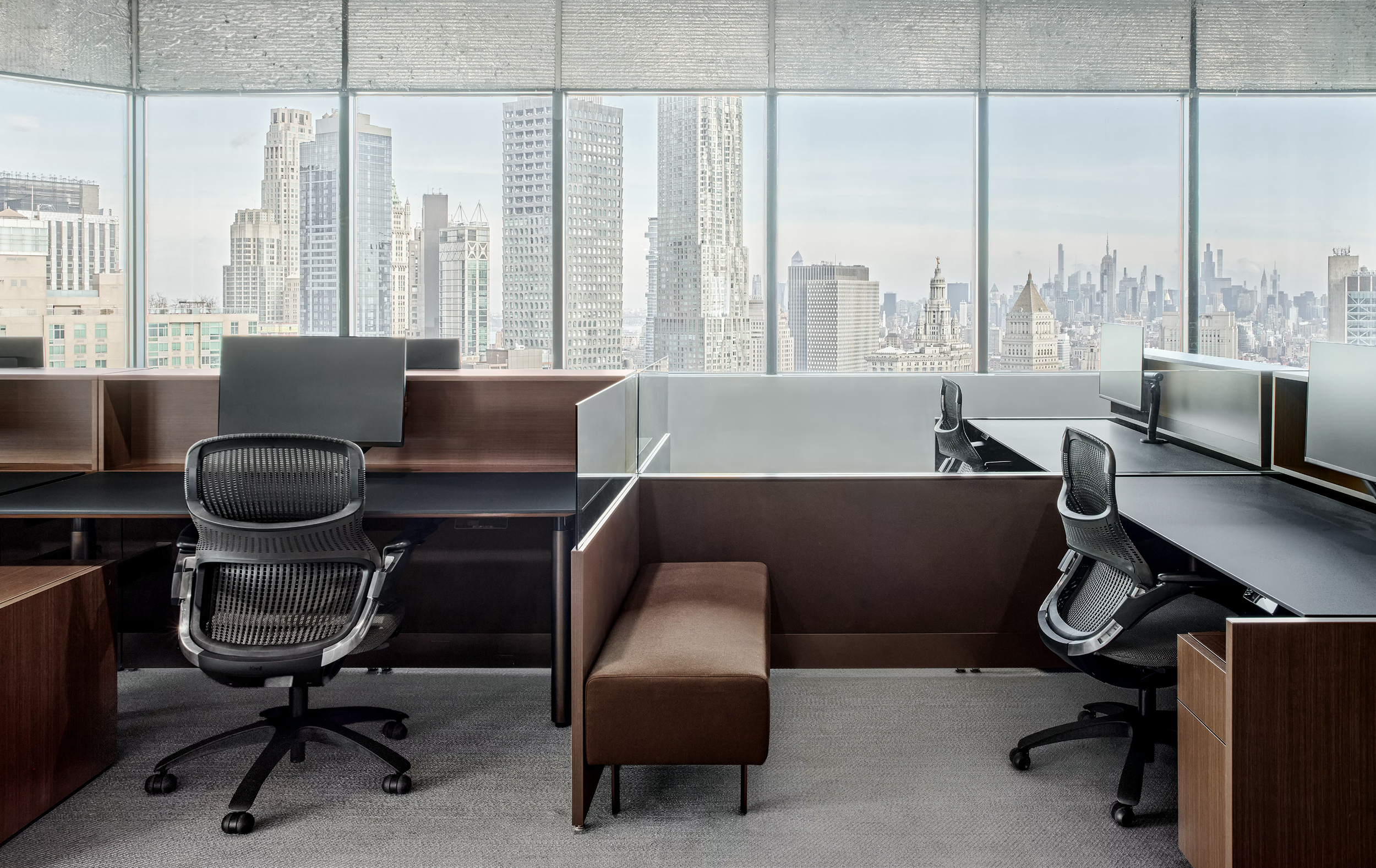 Modern office cubicles with ergonomic chairs, arranged in front of large windows showing a city skyline with tall buildings.