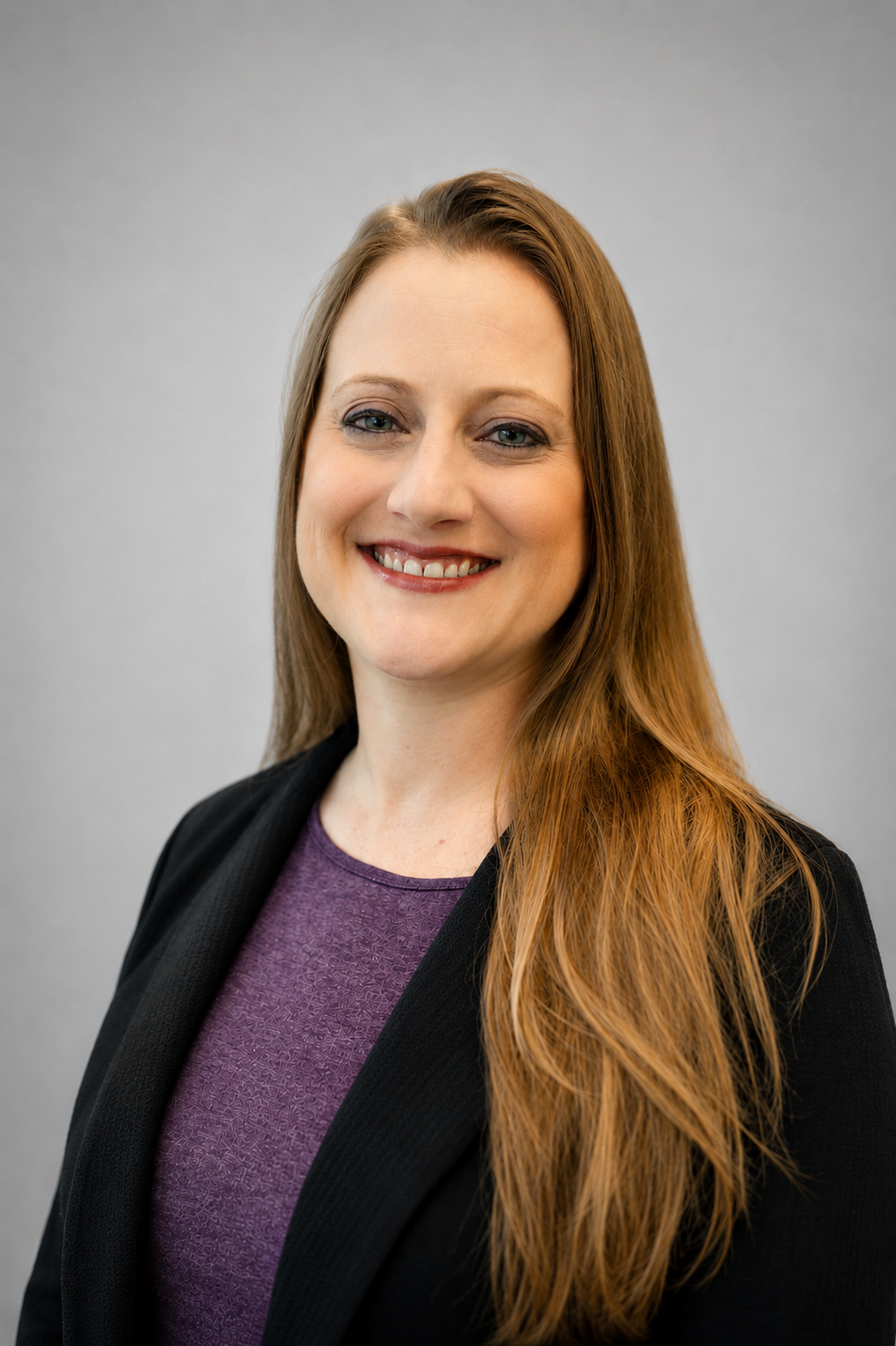A young woman with long reddish-brown hair smiling, wearing a purple top and black blazer, against a plain gray background.