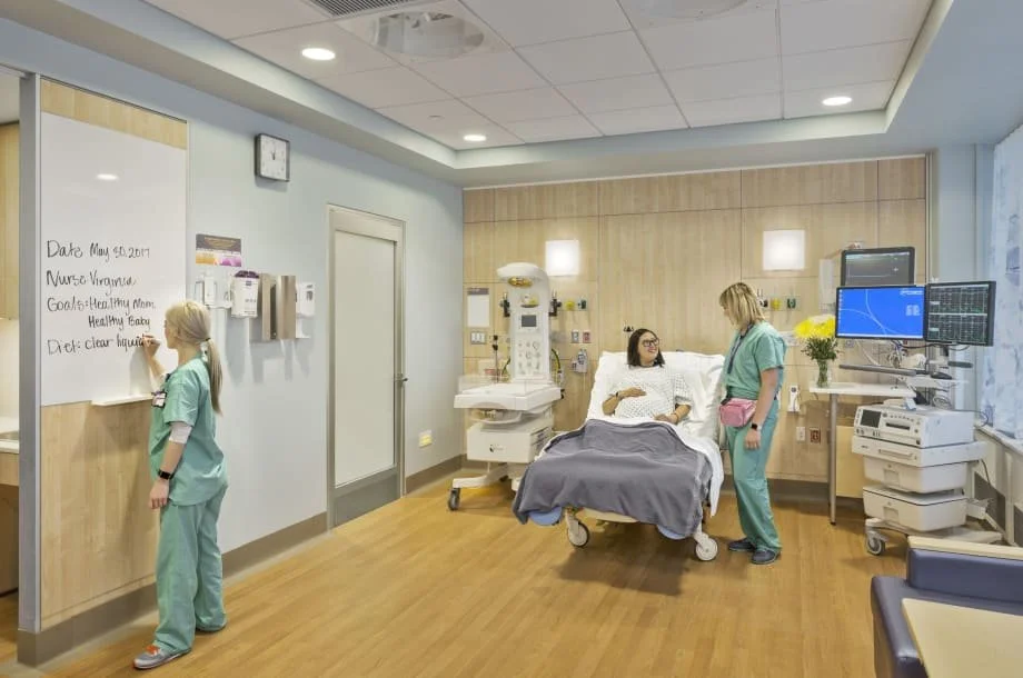 A hospital room with a patient in bed talking to a nurse, a girl in scrubs writing on a whiteboard, and medical equipment around.