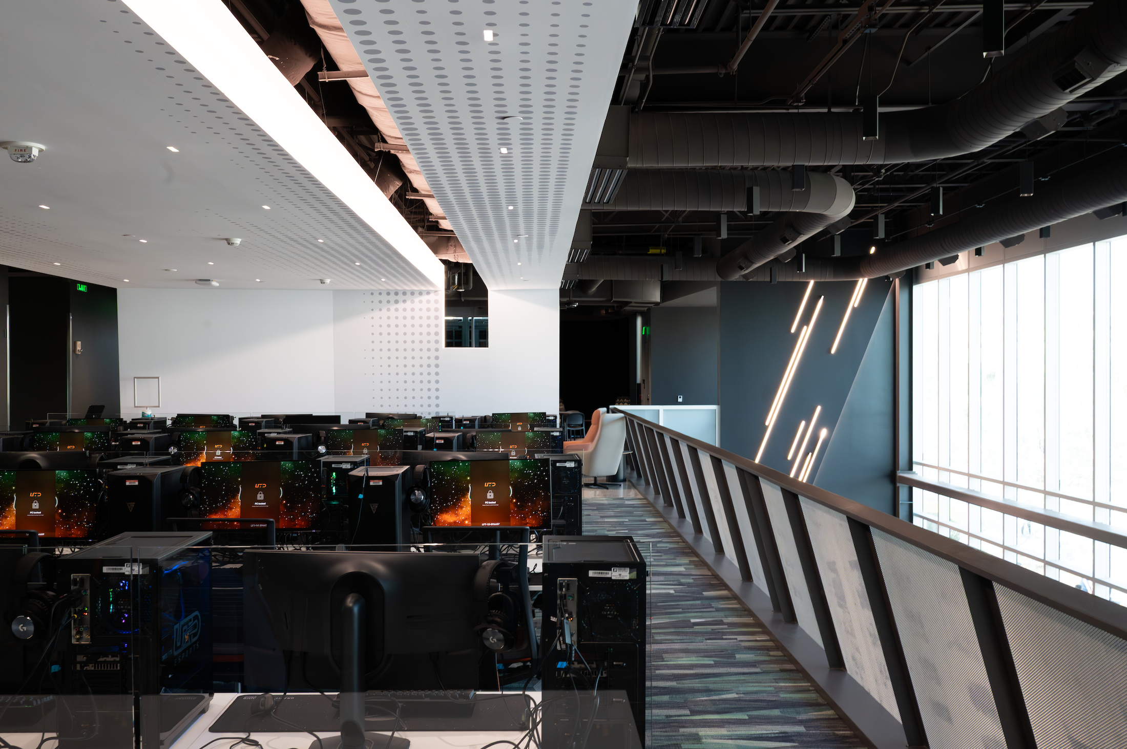 A modern office or computer lab with rows of desktop computers and monitors, large windows on the right side, with ceiling pipes and lighting fixtures visible, and a railing along the right side overlooking the space.