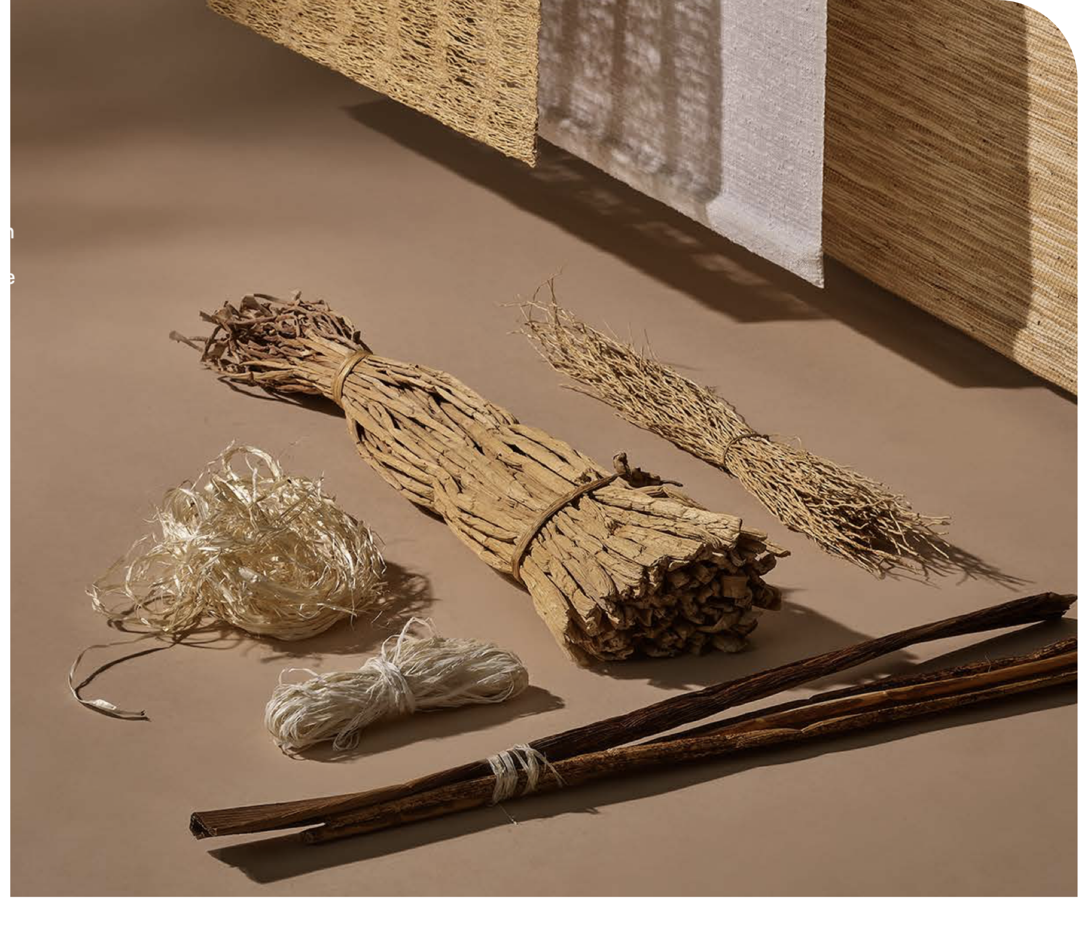 Various natural and handmade objects, including bundles of dried herbs or grasses, and wooden sticks, arranged on a beige surface with a background of stacked mats or panels of different textures and colors.
