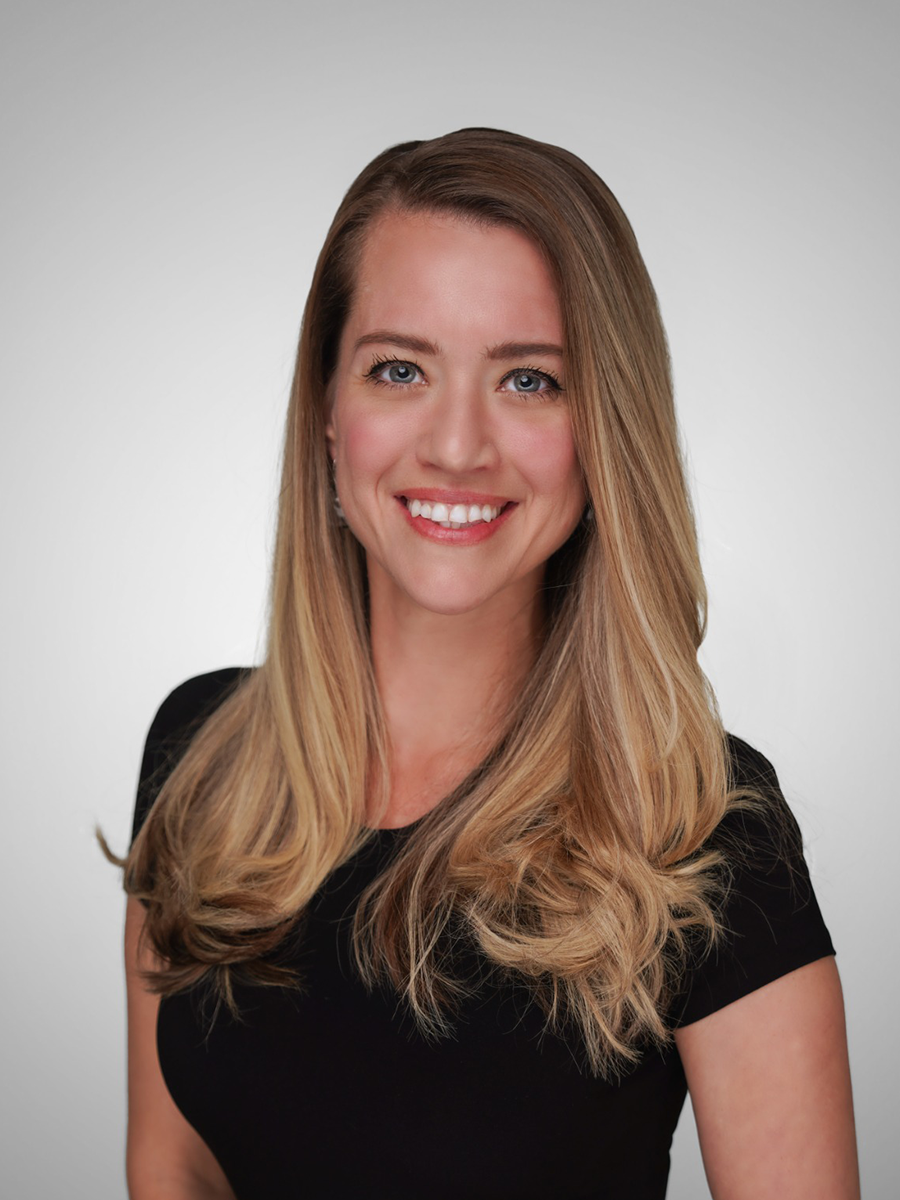 A smiling woman with long, wavy blonde hair wearing a black top, posing against a plain gray background.