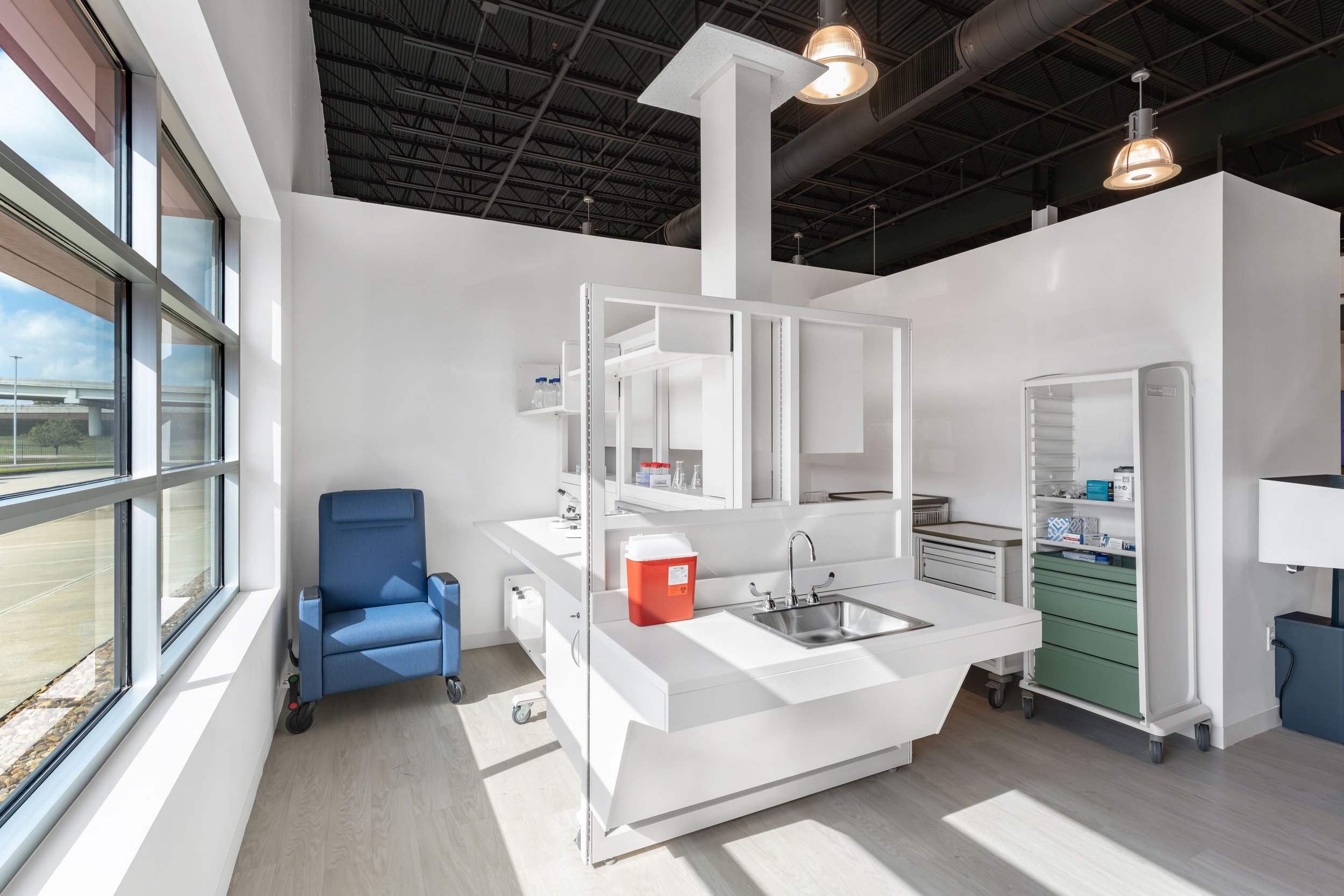 A modern, clean hospital or medical clinic room with white walls and large windows, a blue medical chair, and a small kitchenette area with a stainless steel sink and medical supplies on shelves.