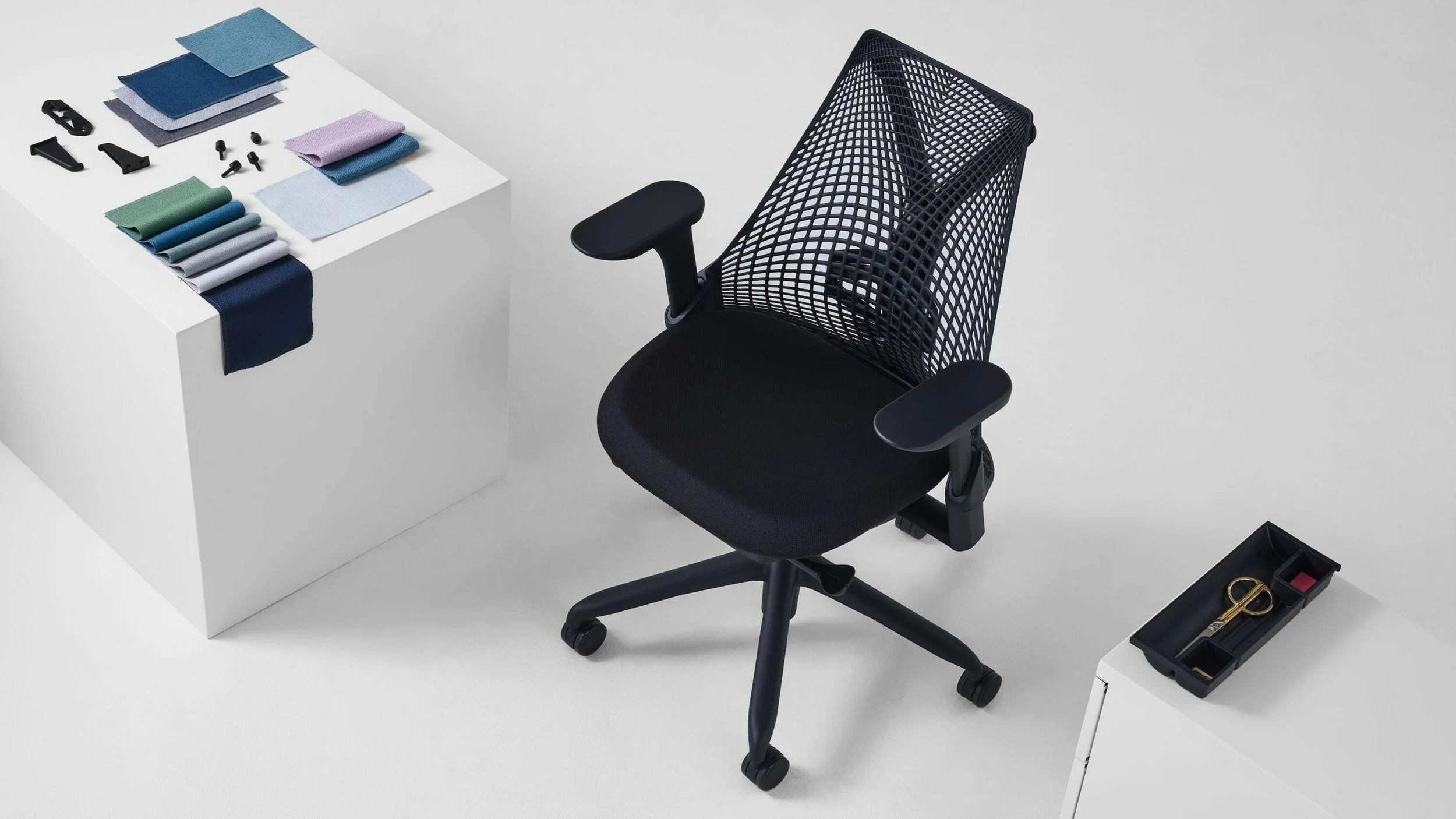 A black ergonomic office chair with a mesh backrest and adjustable armrests positioned next to a white table. The table has fabric samples, screws, and hardware arranged on it, with some fabric swatches hanging off the edge. There is a small black tray with scissors and other tools on a white cabinet nearby.