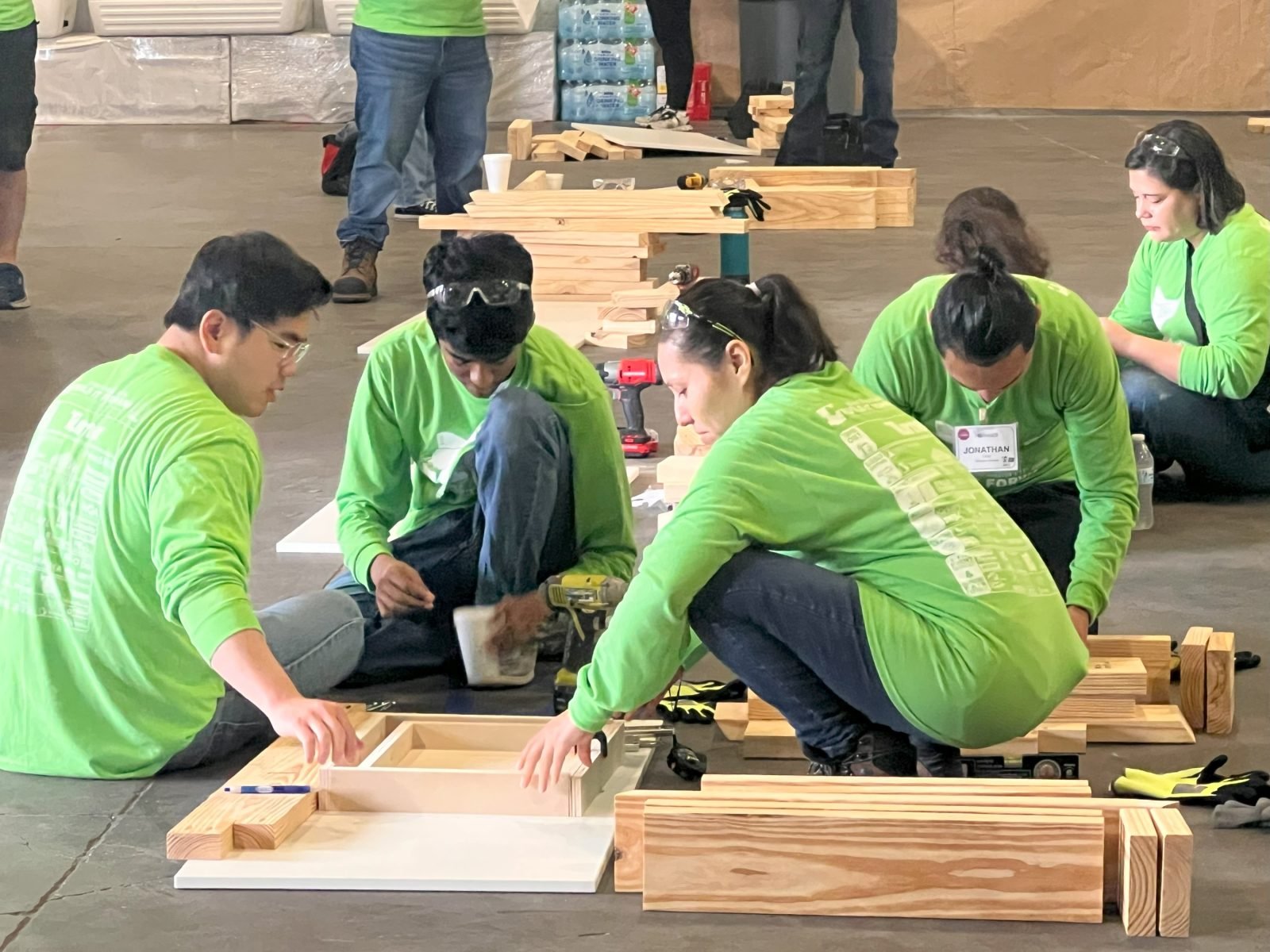 Group of people wearing green shirts working on a woodworking project, assembling furniture with wood pieces on the floor inside a workshop or event space.