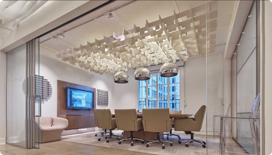 Modern conference room with beige chairs around a table, wall-mounted TV, and unique ceiling design with hanging light fixtures.